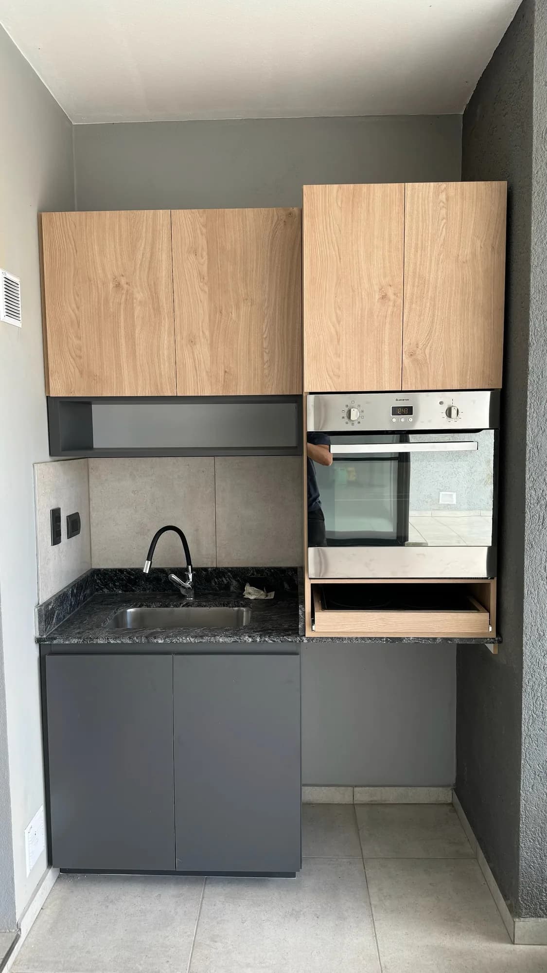Cocina compacta con alacenas en melamina símil roble, nicho abierto gris, horno empotrado en columna de madera, mesada de granito oscuro con bacha de acero y bajomesada antracita; toma frontal