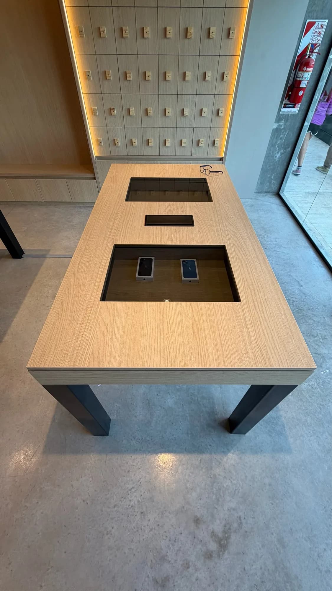 Mesa de exhibición en chapa de roble con patas negras y dos huecos vidriados con teléfonos, frente a un panel de casilleros iluminado; toma cenital en un local