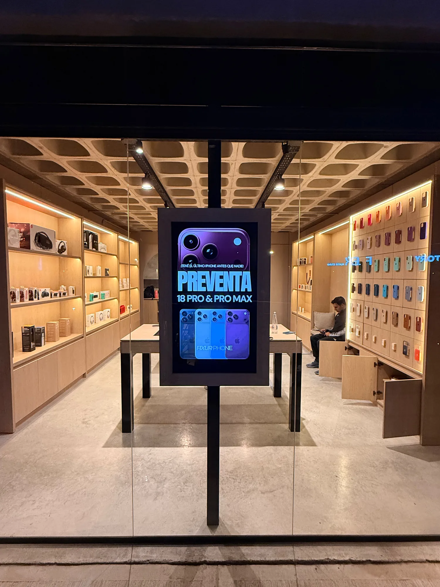 Local de tecnología visto desde la vitrina de noche: estantes y nichos de melamina símil roble con tiras LED cálidas, panel de casilleros con fundas de colores, mesa de exhibición central y pantalla digital