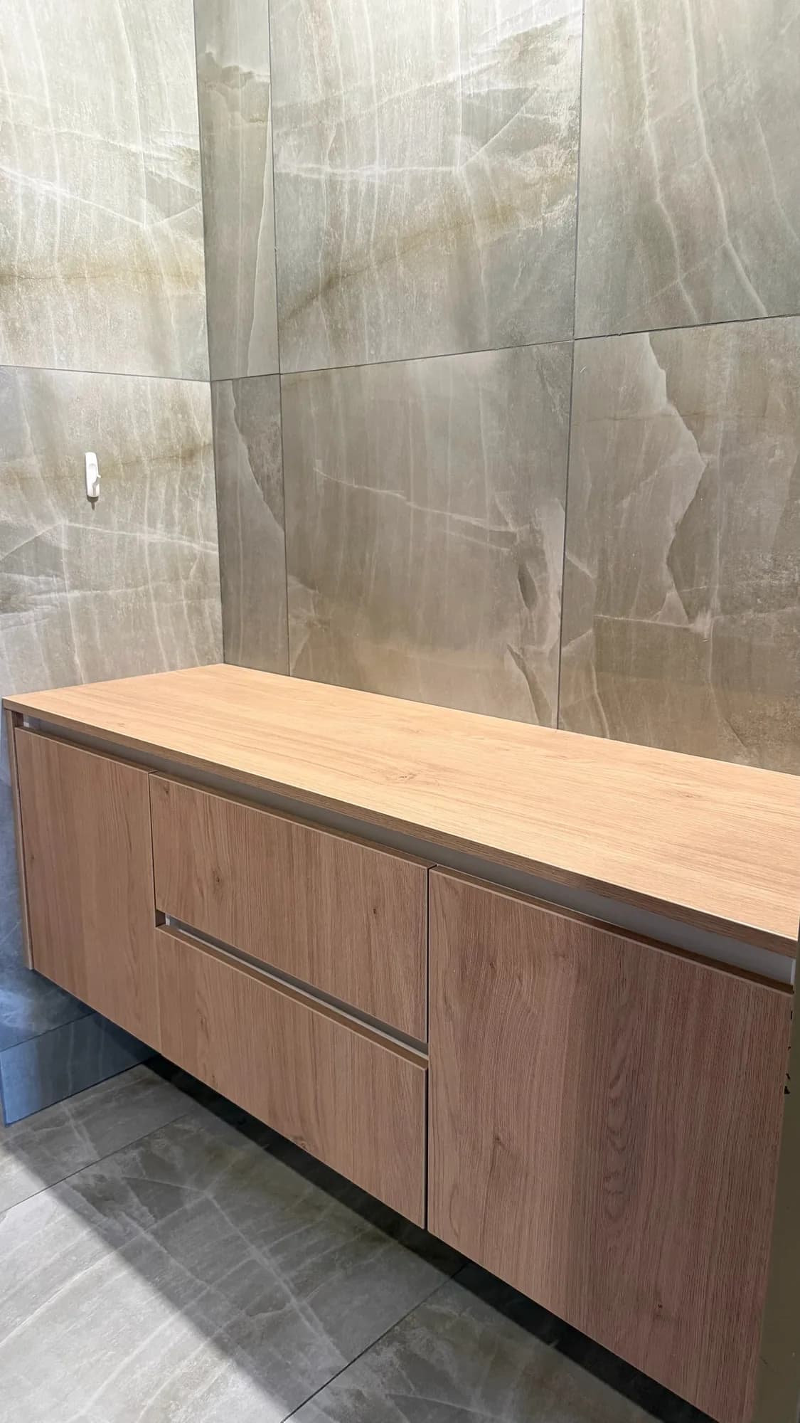 Vanitory suspendido de melamina símil roble con dos cajones y una puerta, sobre pared de porcelanato símil ónix; toma lateral en ángulo