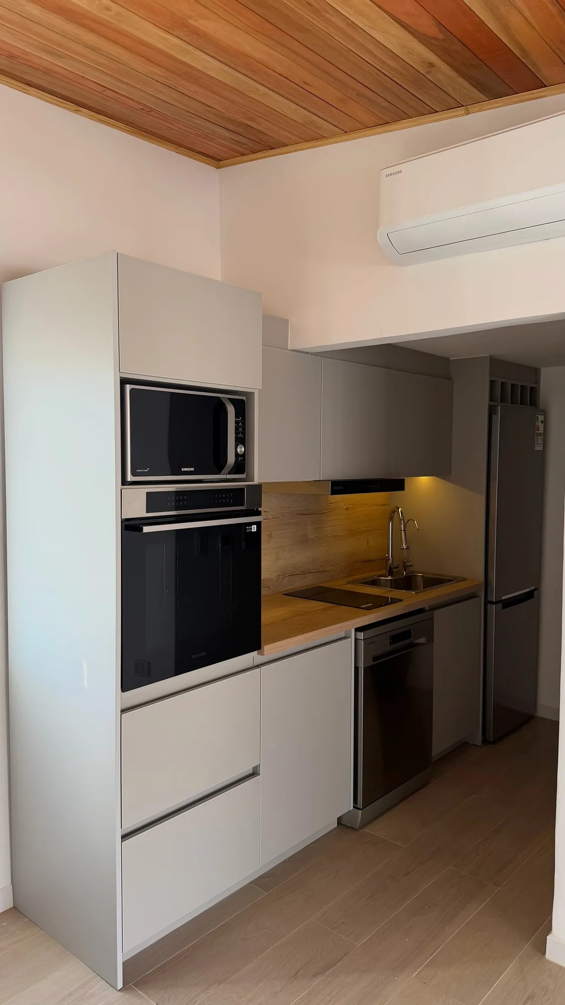 Cocina lineal compacta en gris claro mate con columna de horno y microondas, mesada símil madera y heladera al fondo, bajo cielorraso de listones; toma en diagonal