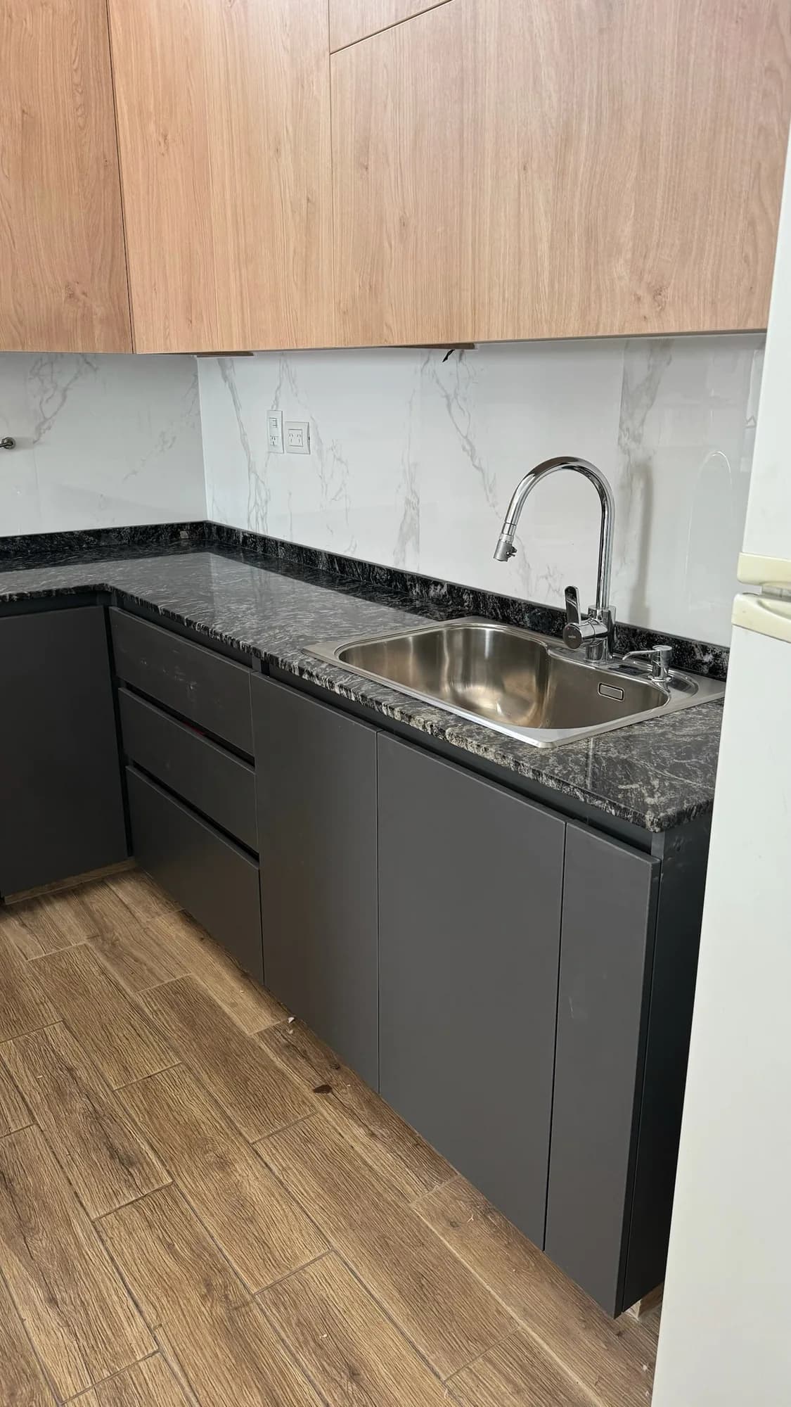 Rincón de cocina con bajomesadas antracita sin tiradores, cajonera, mesada de granito negro con bacha de acero y grifería extraíble, alacenas símil roble y revestimiento símil mármol