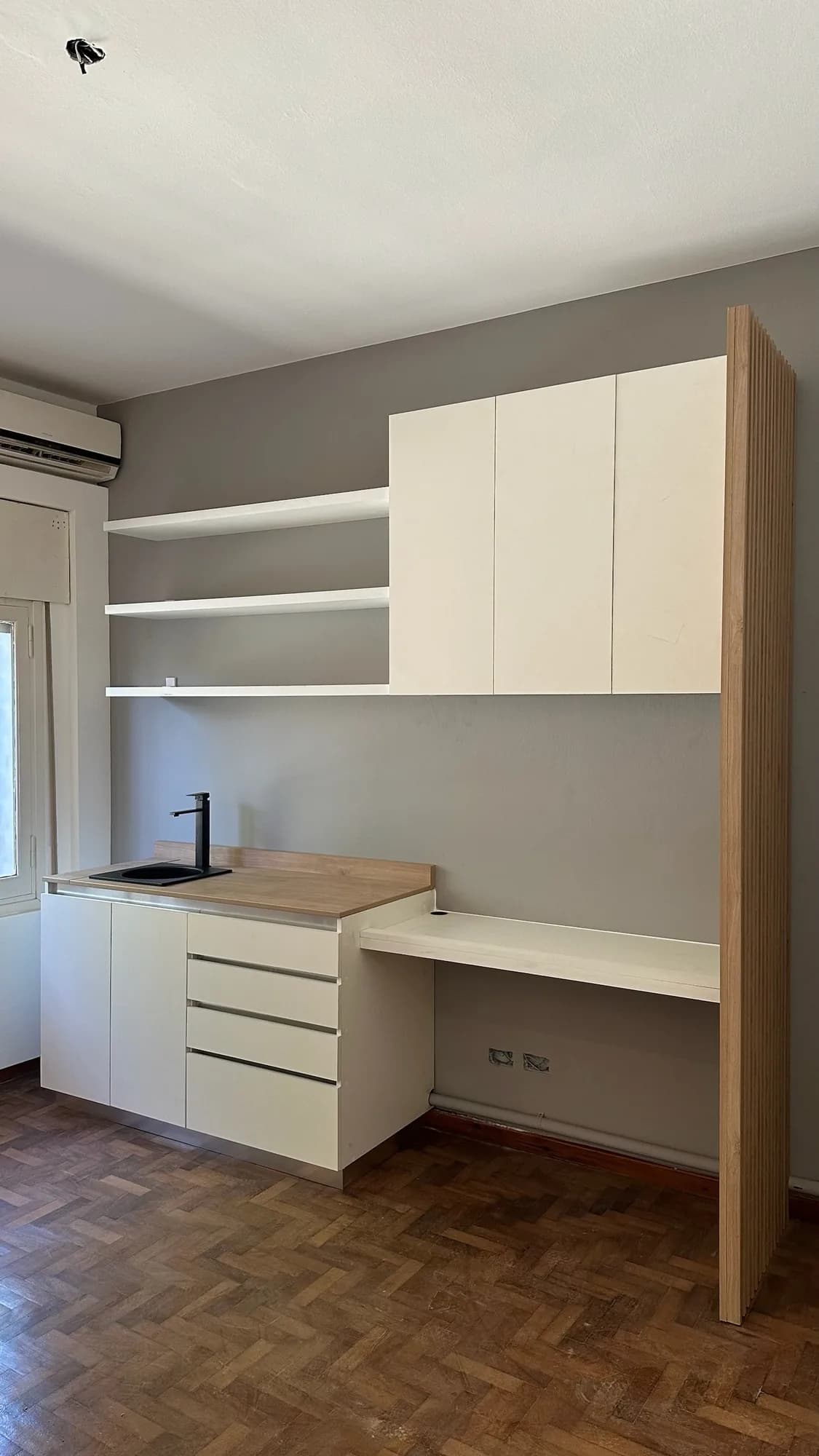 Kitchenette con escritorio flotante, alacenas altas blancas, repisas y panel de listones de madera clara en el lateral; toma frontal sobre pared gris