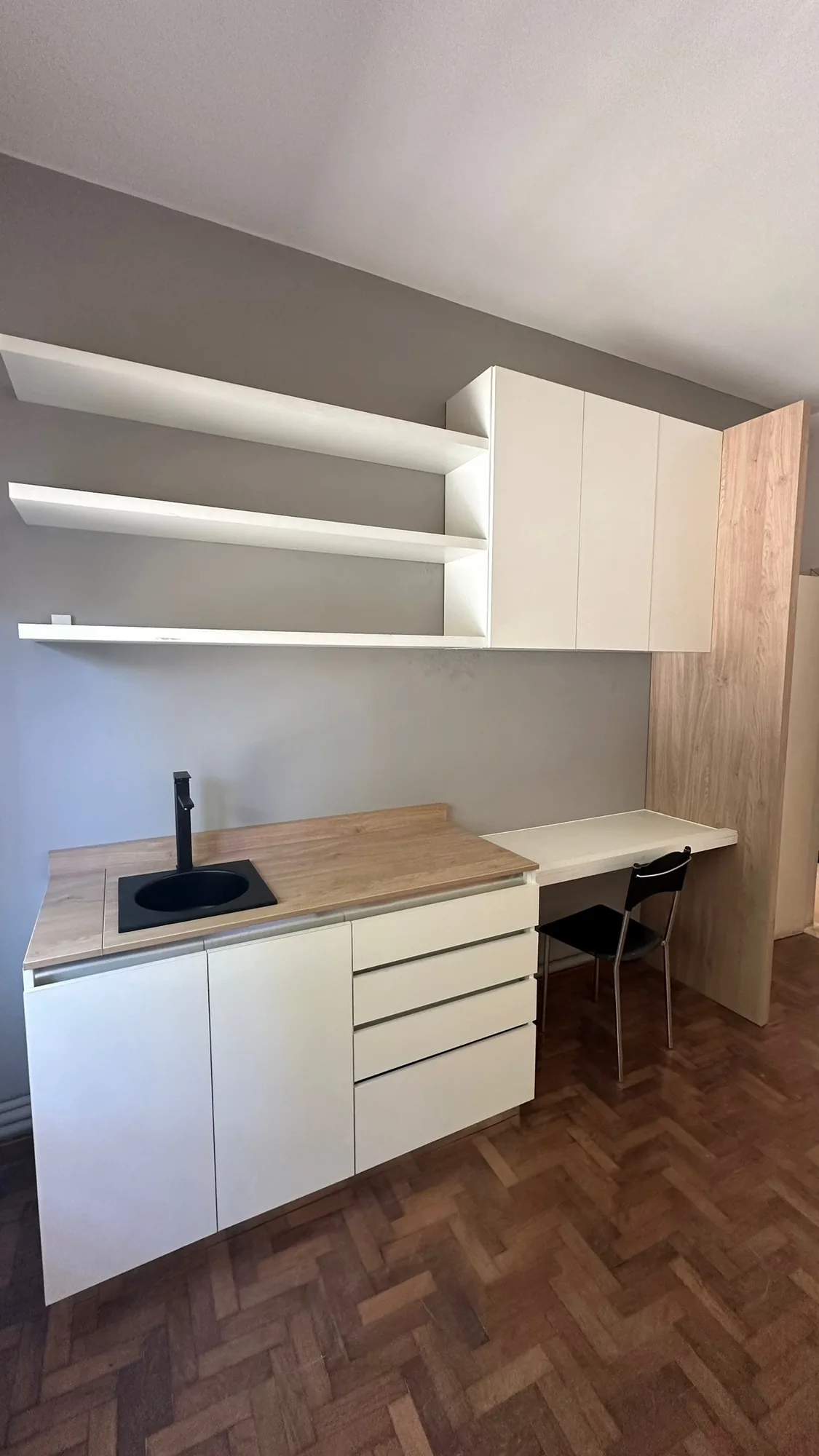 Kitchenette en melamina blanca con bacha negra, mesada símil madera, repisas flotantes, alacenas altas y escritorio integrado junto a un lateral símil roble; toma frontal general