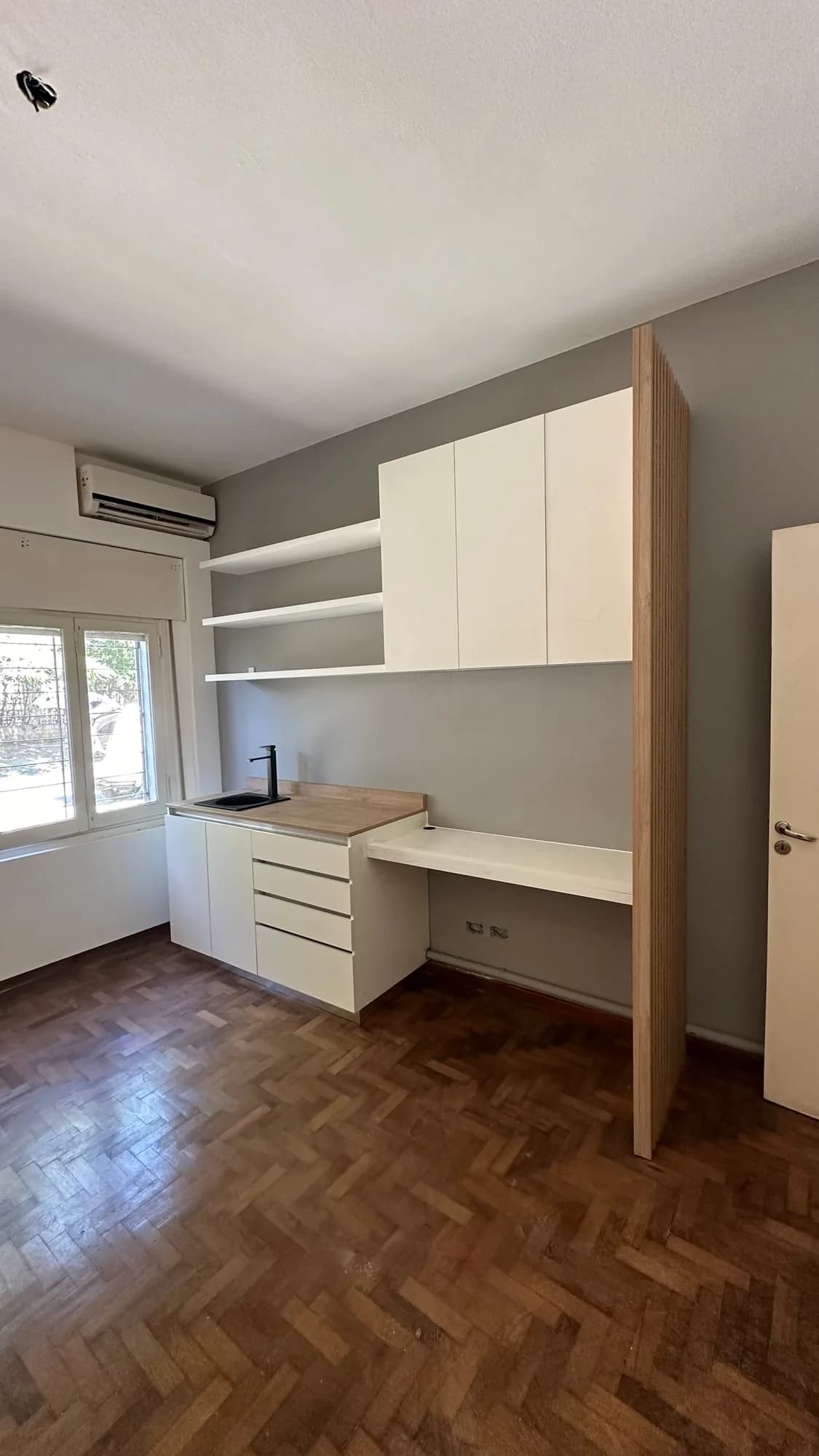 Ambiente con kitchenette integrada en pared gris, con alacenas blancas, repisas flotantes, escritorio y panel lateral de listones de madera clara; toma general amplia