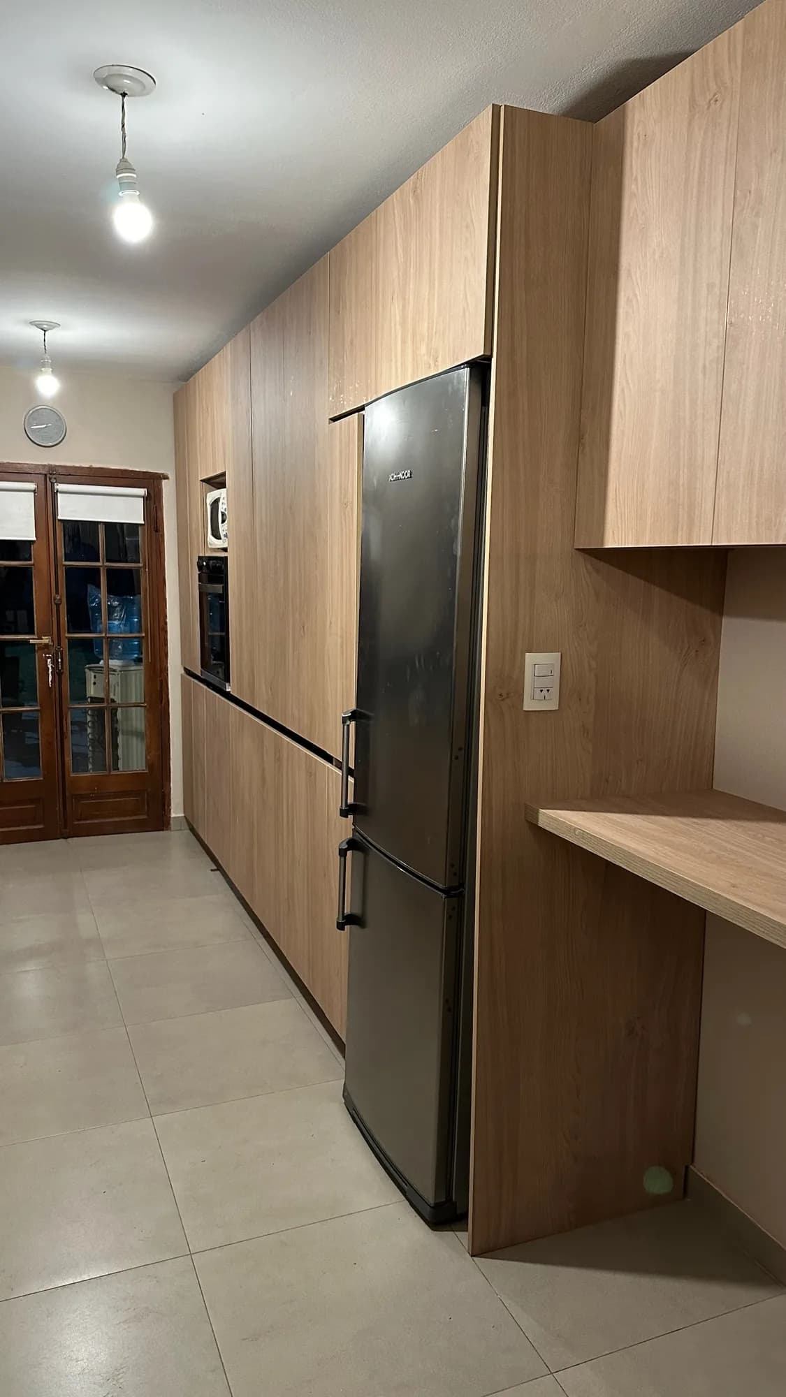 Cocina en línea con columnas de melamina símil roble hasta el techo, heladera exenta de acero y barra auxiliar; toma en diagonal desde el fondo