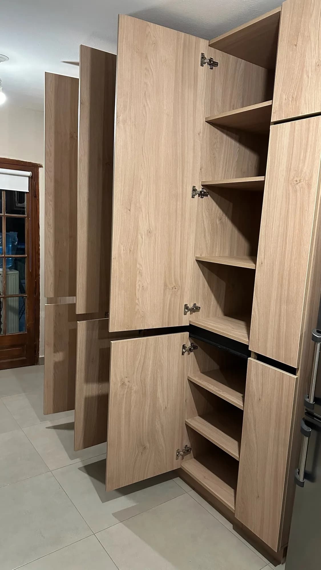 Columna de cocina con puertas abiertas que muestran estantes y bisagras en melamina símil roble; toma en diagonal junto a una puerta vidriada