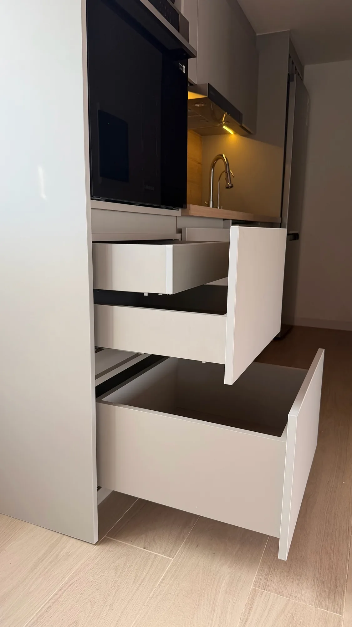 Columna de cocina con tres cajones abiertos de frentes lisos y cuerpos de melamina gris, con horno, grifo y mesada iluminados al fondo; toma baja en diagonal