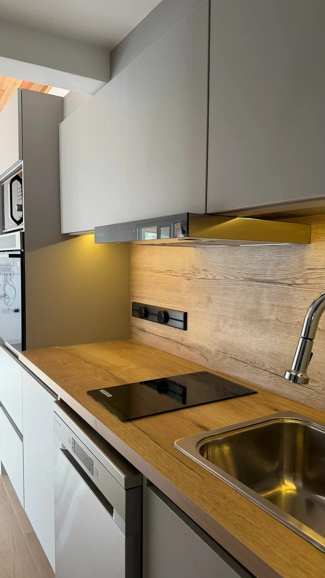 Cocina lineal con alacenas altas grises sin tirador, campana, panel de laminado símil roble con tomas y mesada con anafe, bacha y lavavajillas. Toma en ángulo desde la entrada