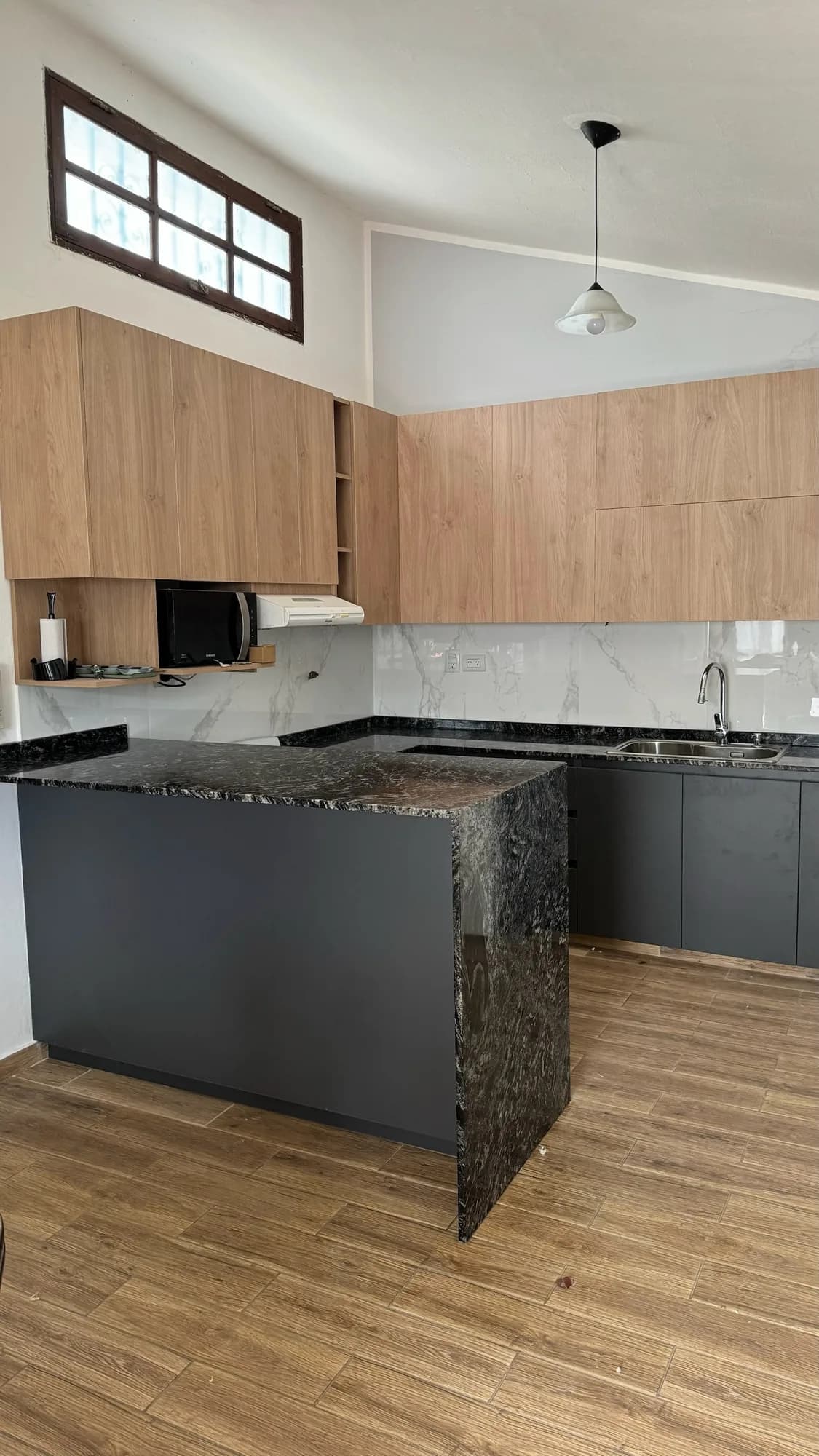 Cocina en U con península de granito negro en cascada, bajomesadas gris antracita y alacenas en melamina símil roble; revestimiento símil mármol y piso símil madera; toma general