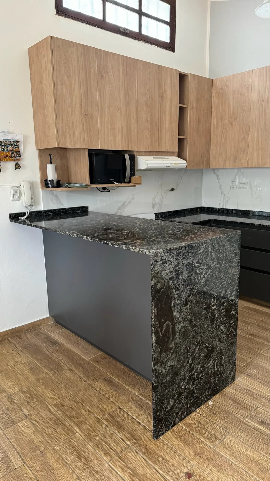 Península de cocina con mesada de granito negro en cascada, frente gris antracita y alacenas en melamina símil roble con microondas en repisa abierta; toma en diagonal