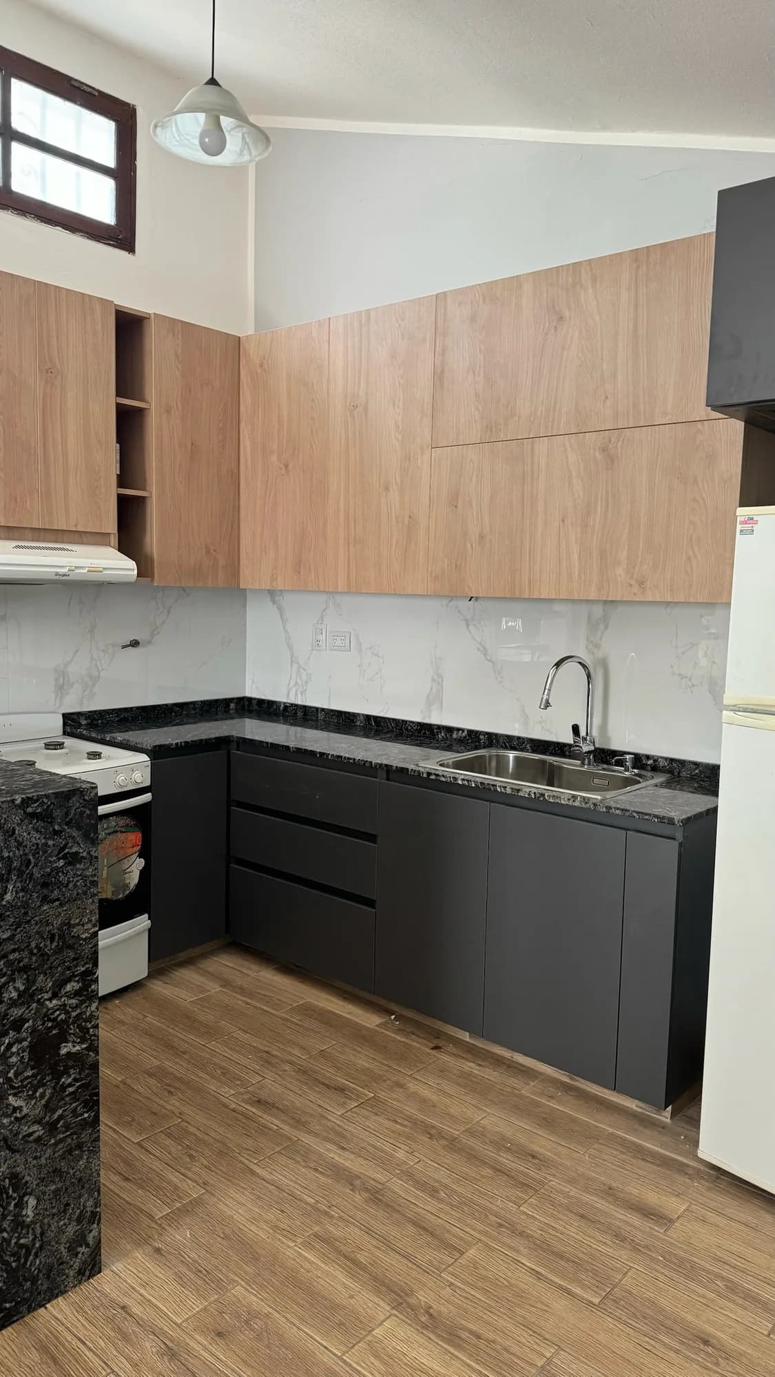 Cocina en L con bajomesadas antracita, mesada de granito negro con bacha de acero, alacenas hasta el techo en melamina símil roble y revestimiento símil mármol; toma en diagonal