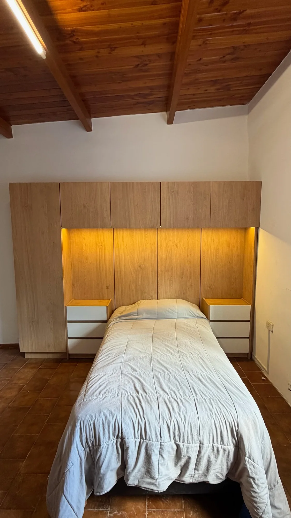 Dormitorio con cabecera de placard y alacenas en melamina símil roble, nichos con LED y mesas de luz de cajones blancos a ambos lados de la cama; toma frontal bajo techo de madera