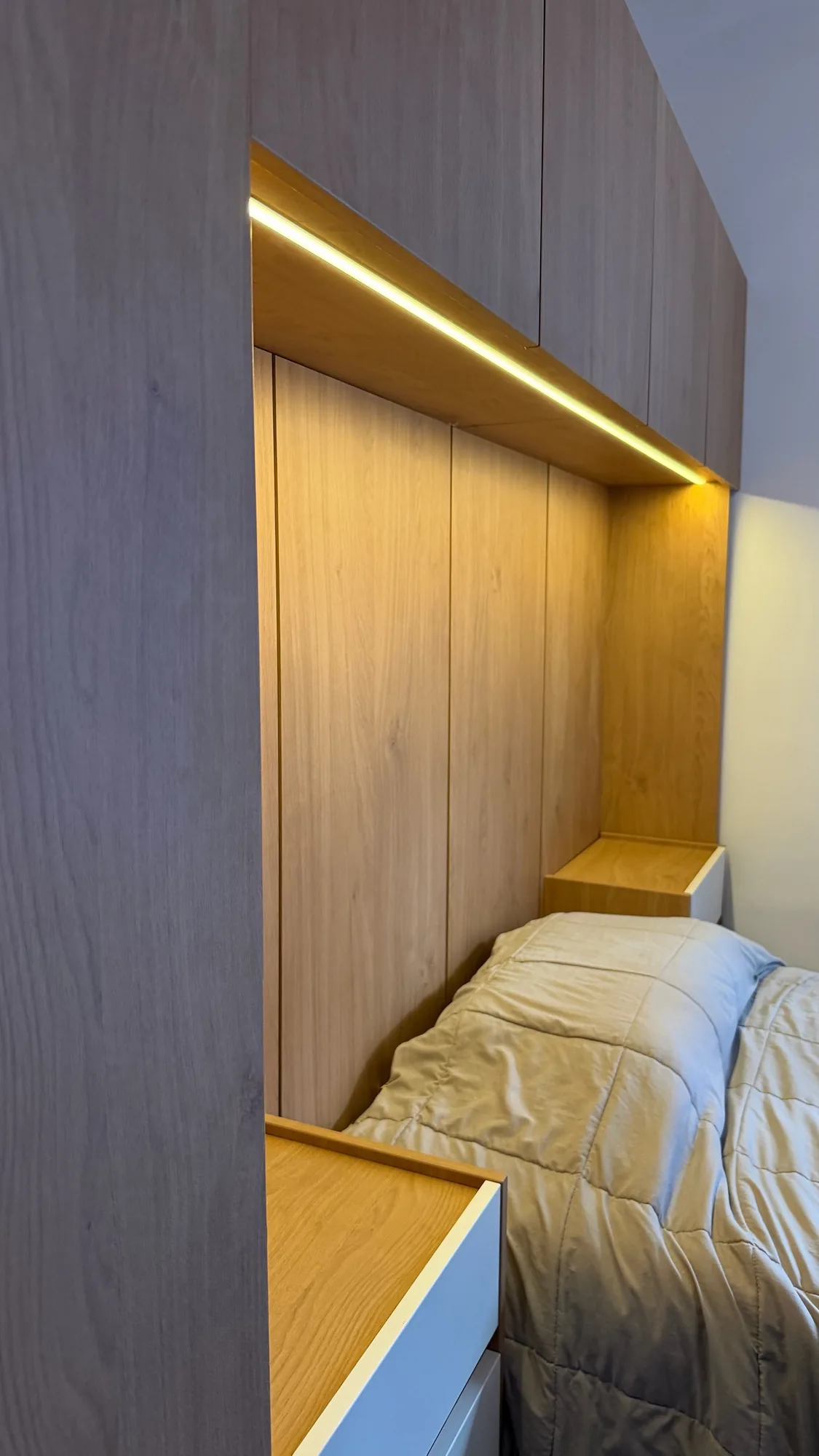 Cabecera de dormitorio con paneles símil roble, alacenas superiores y tira LED cálida bajo ellas, con mesas de luz blancas junto a la cama; toma lateral en ángulo