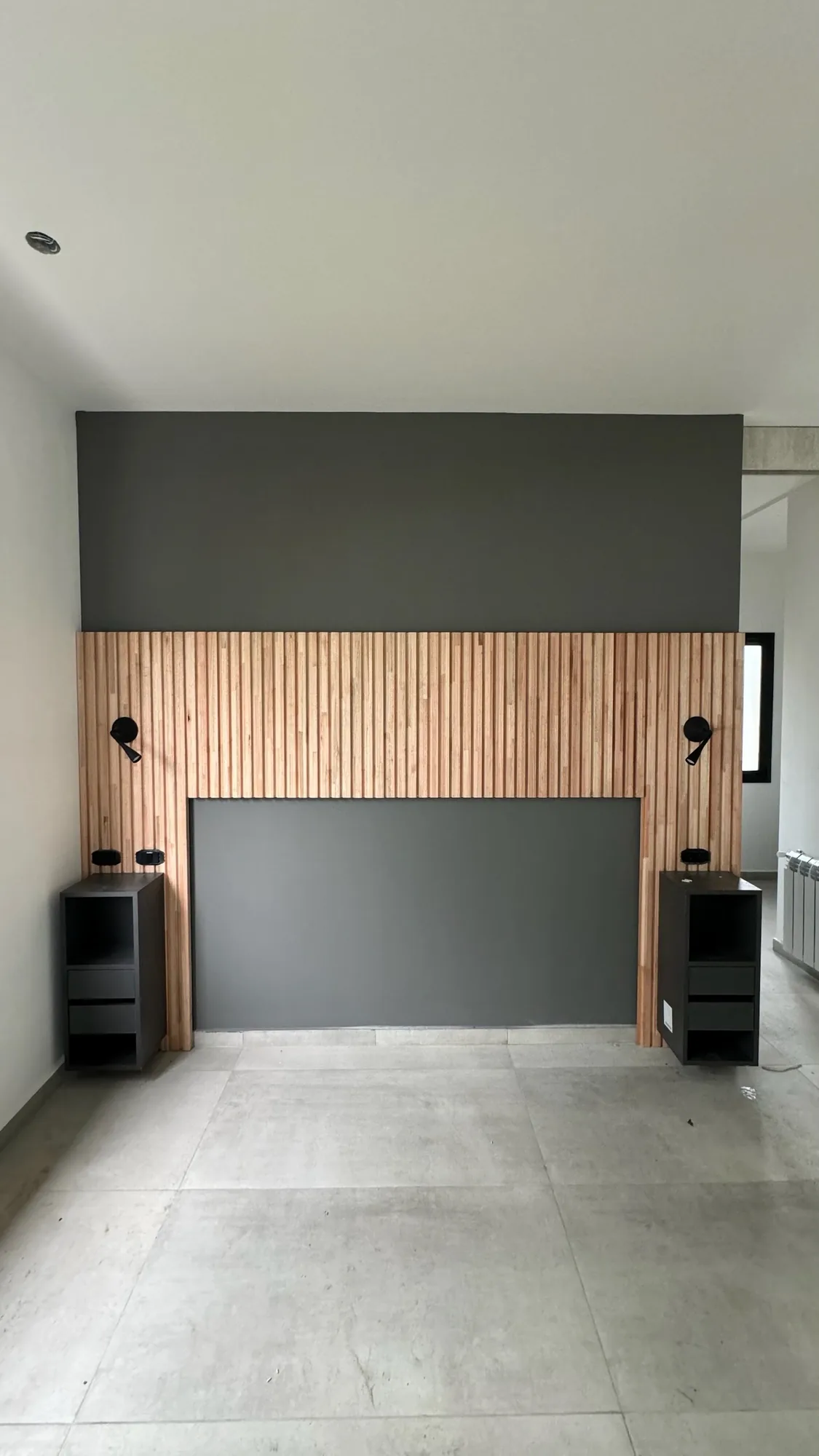 Cabecera de dormitorio con panel de listones de madera natural sobre pared gris oscura, apliques de lectura y dos mesas de luz flotantes negras con cajones; toma frontal simétrica