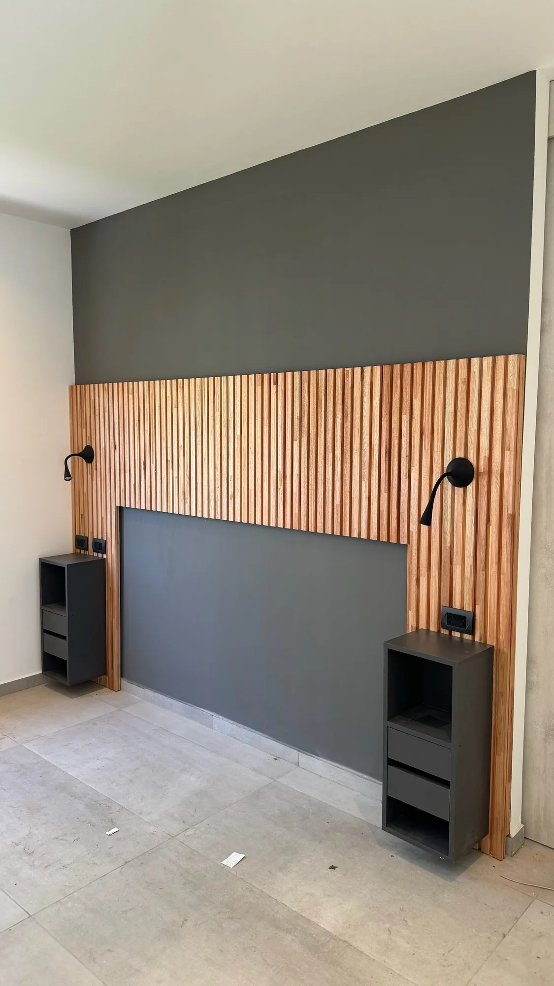 Cabecera de dormitorio con panel de listones de madera natural sobre pared gris oscura, apliques de lectura y dos mesas de luz flotantes negras con cajones; toma angulada desde la izquierda