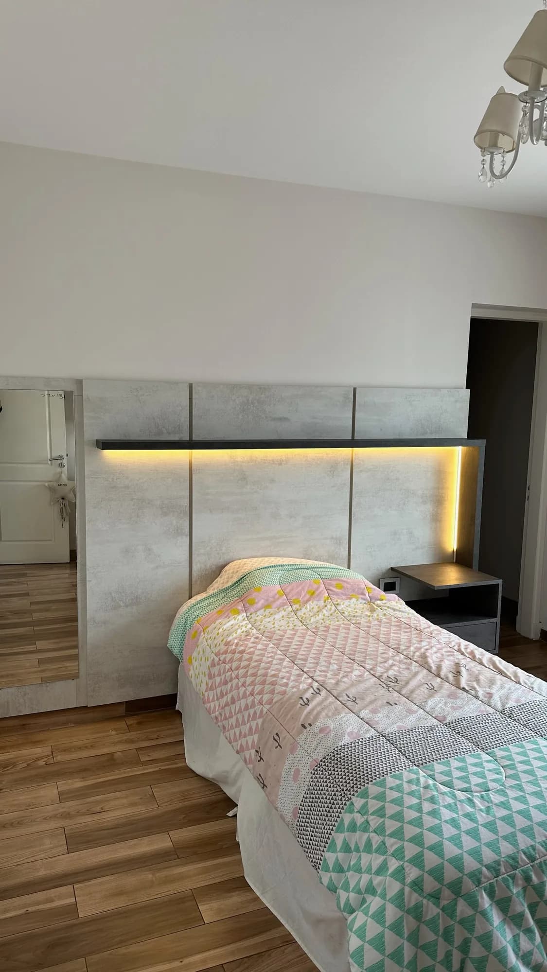 Respaldo de cama en paneles símil cemento con repisa superior y tira LED cálida, mesa de luz suspendida oscura y espejo lateral; toma frontal del cabecero