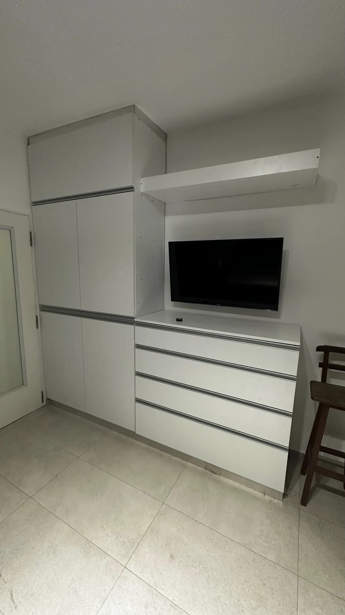 Placard alto blanco de cuatro puertas junto a una cómoda de cuatro cajones con TV en la pared y estante flotante blanco; toma en diagonal en un ambiente con piso de porcelanato