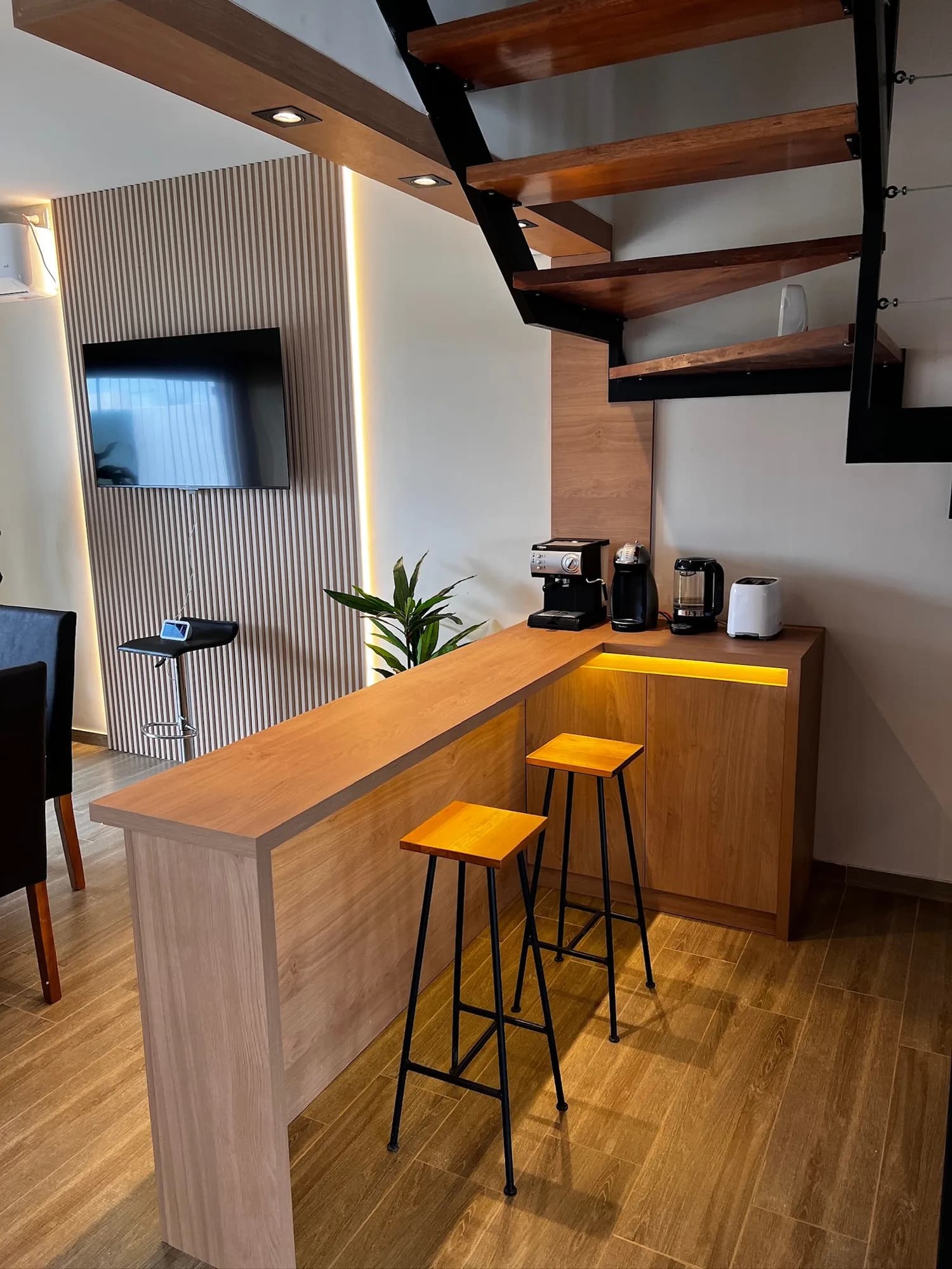 Barra de desayuno en L con tapa de melamina símil madera, mueble bajo con tira LED y dos banquetas de hierro, bajo una escalera abierta con spots; a la izquierda, TV sobre panel de listones. Toma vertical