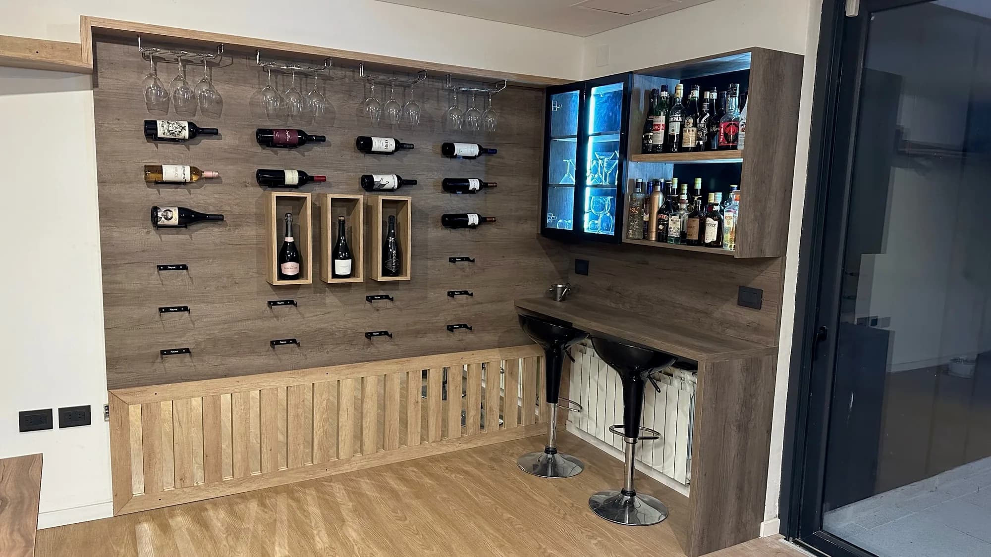 Rincón bar con panel de melamina símil madera oscura para botellas y copas colgadas, nichos de madera, vitrina con luz LED azul, barra con dos banquetas y cubreradiador de listones; toma frontal en ángulo