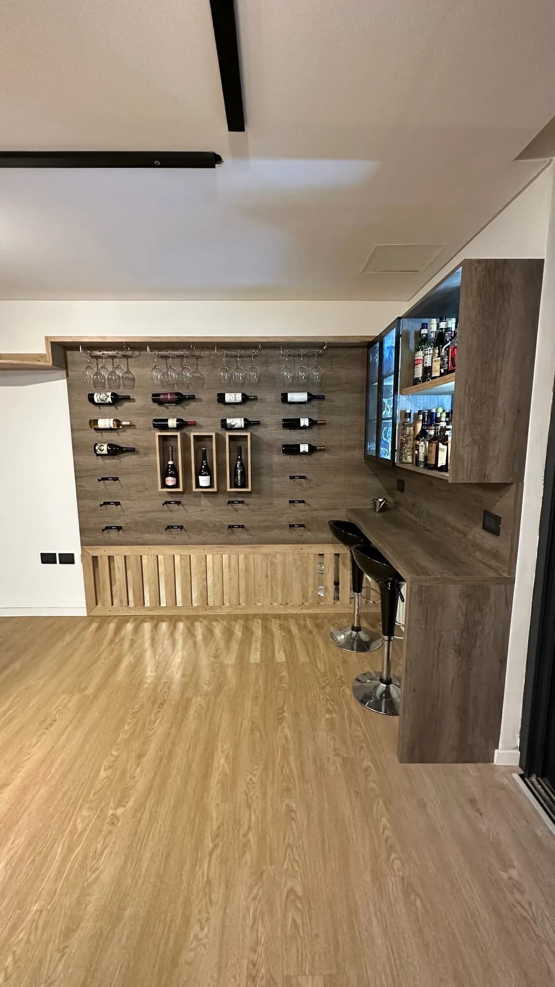 Vista amplia del rincón bar con panel de botellas, vitrinas y barra con banquetas, sobre un piso símil madera; toma vertical desde lejos