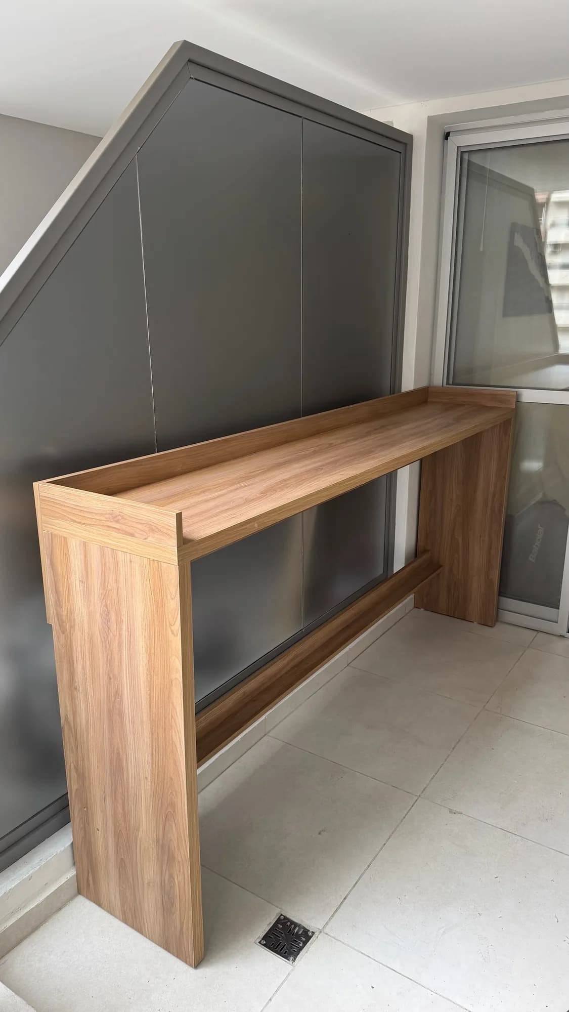 Mesa alta tipo barra en melamina símil nogal con travesaño apoya pies, junto a un placard gris con frentes lisos que sigue la pendiente de la escalera. Toma en ángulo en un balcón con porcelanato claro