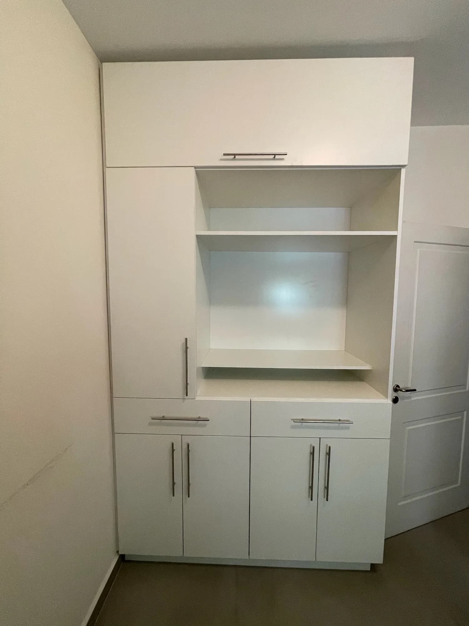 Mueble alto de melamina blanca con alacena superior, nicho abierto con estantes, puertas y cajones inferiores con tiradores de barra; toma frontal en una habitación vacía