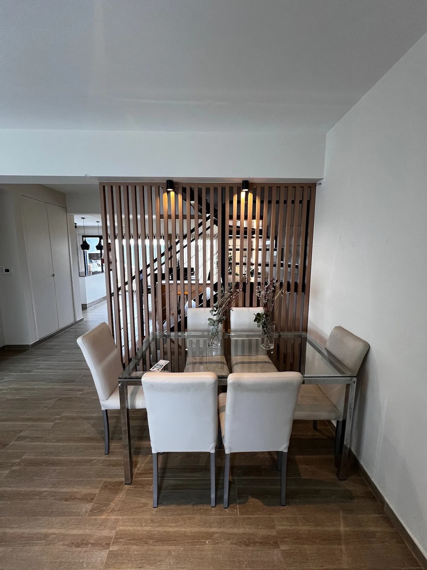 Divisor de ambientes de listones verticales de madera con dos apliques negros, detrás de una mesa de vidrio con seis sillas claras; al fondo se ve la escalera y la cocina. Toma frontal vertical
