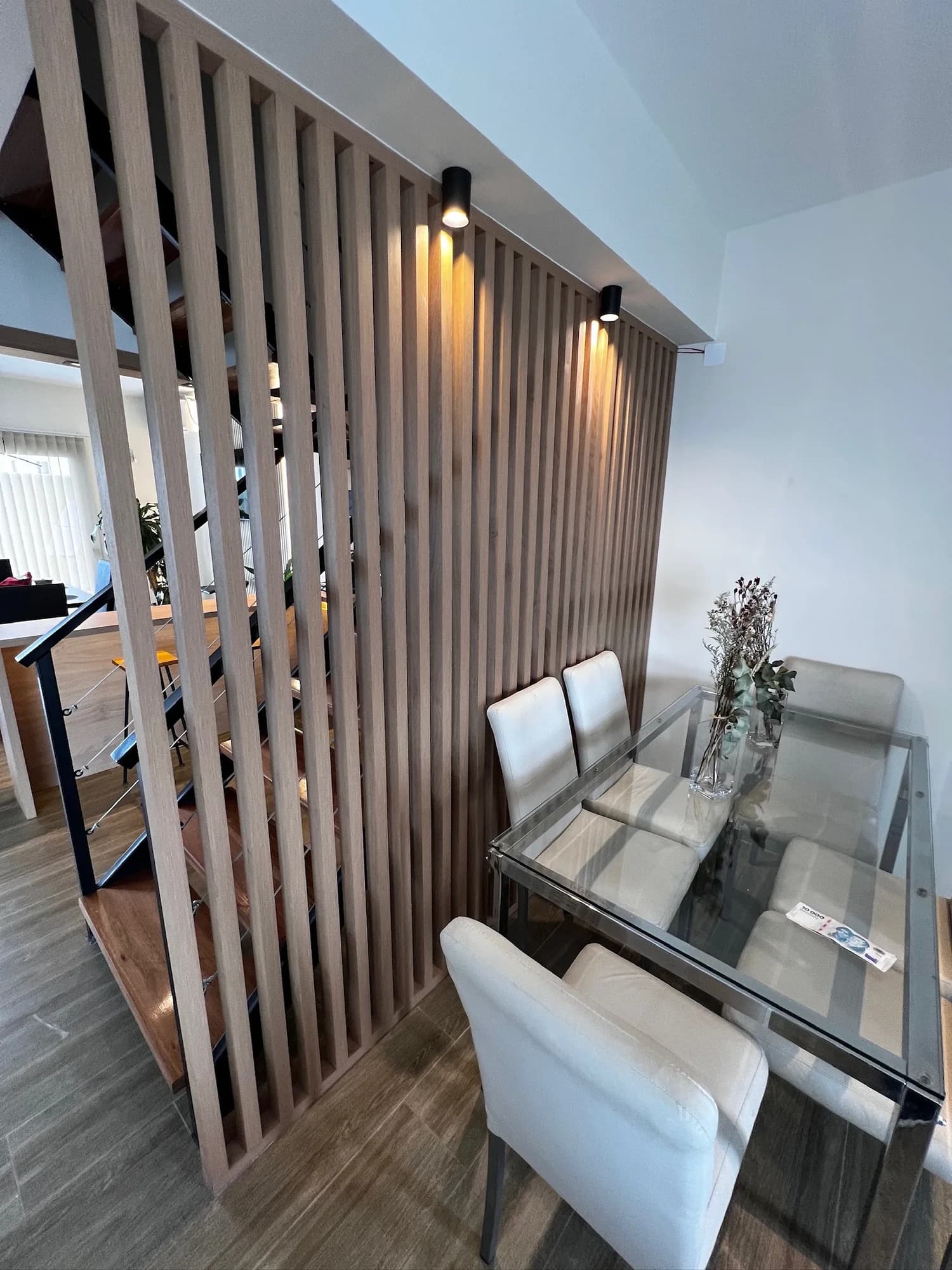 Divisor de listones verticales de madera con apliques negros visto en diagonal, junto a una mesa de vidrio con sillas blancas; a la izquierda se asoma la escalera con barandas de cable