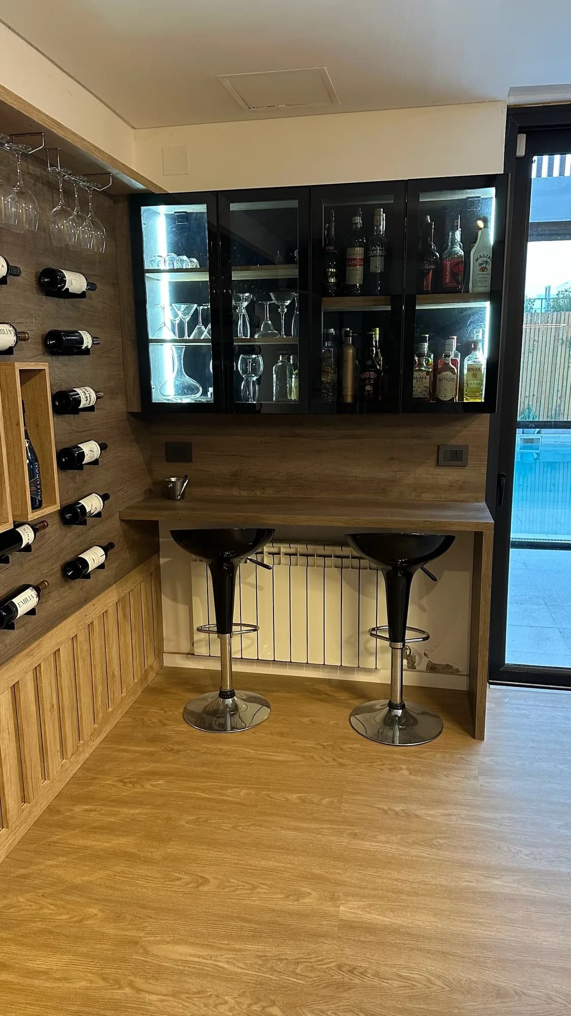 Vitrina de cuatro puertas de vidrio con LED sobre una barra de melamina símil madera con dos banquetas negras; a la izquierda, panel de botellas; toma frontal