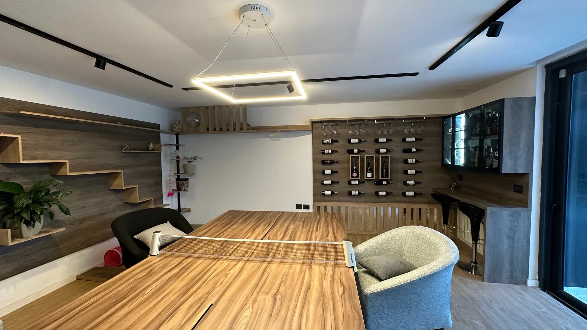 Sala de juegos con panel de botellas, vitrinas y barra con banquetas a la derecha, repisas escalonadas a la izquierda y lámpara LED colgante sobre una mesa de ping pong; toma general