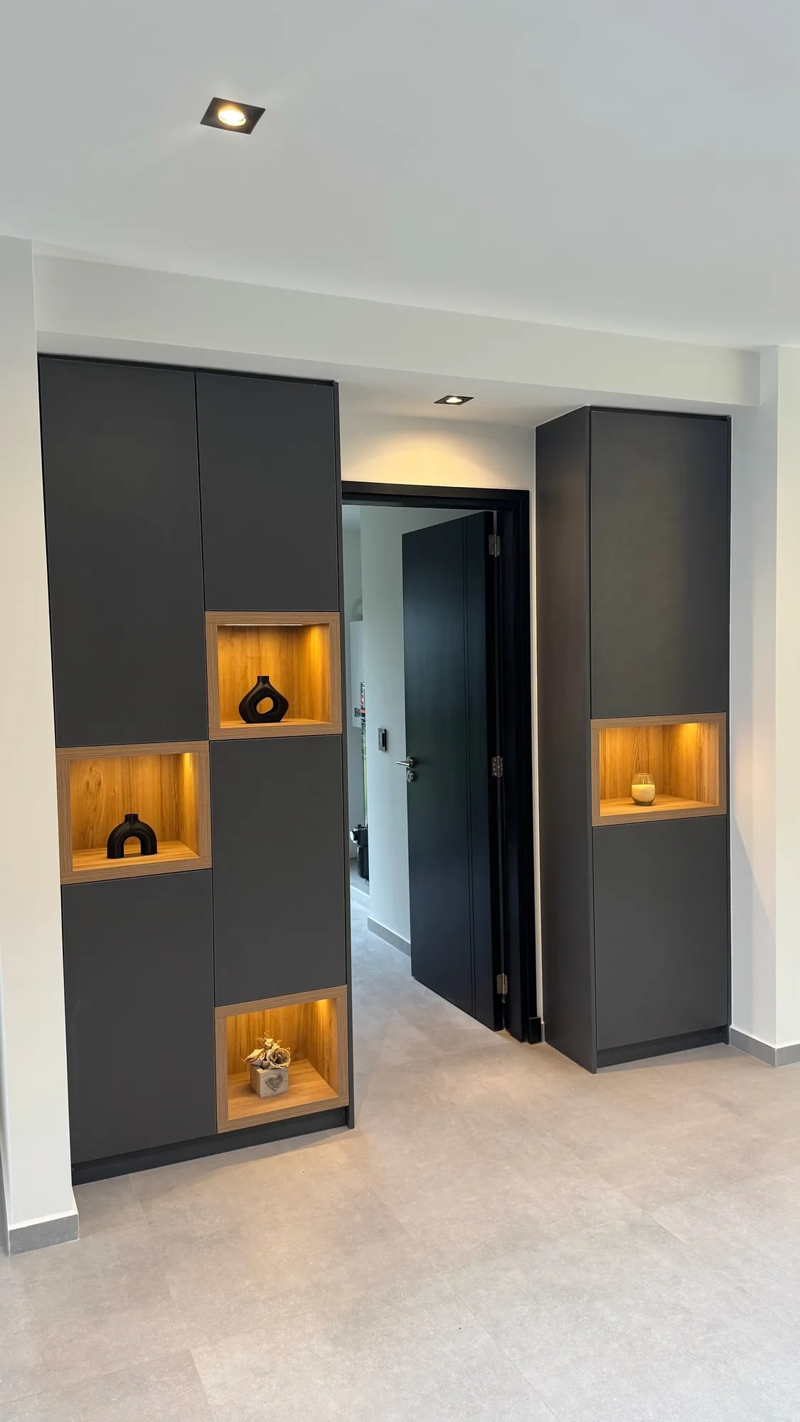 Muebles altos de recibidor en melamina gris grafito sin tirador con nichos de madera iluminados con LED cálido y piezas decorativas, a ambos lados de una puerta oscura. Toma frontal con spots empotrados en el techo