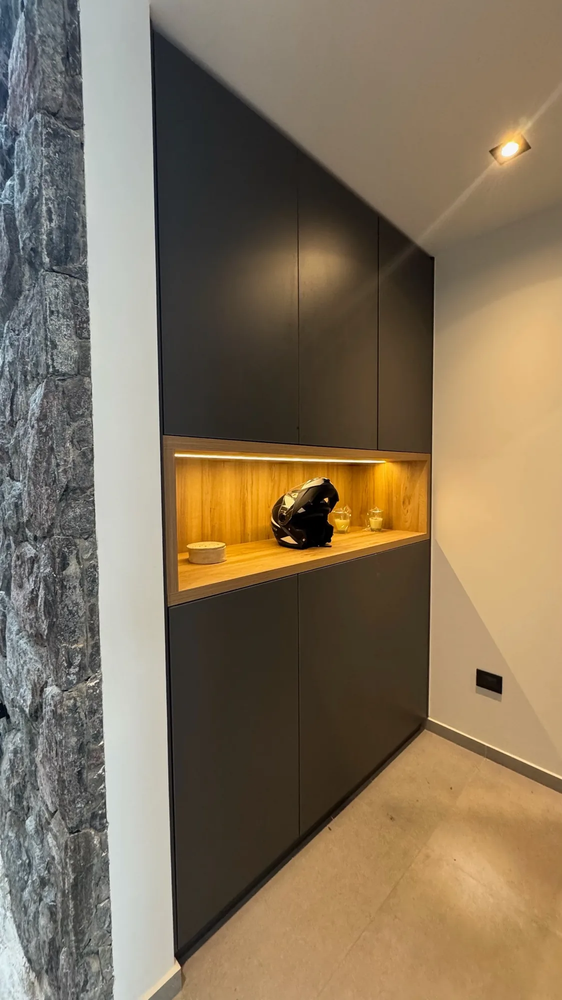 Mueble alto de frentes lisos gris oscuro con nicho central de madera clara iluminado con tira LED, junto a una pared de piedra; toma vertical frontal en un acceso