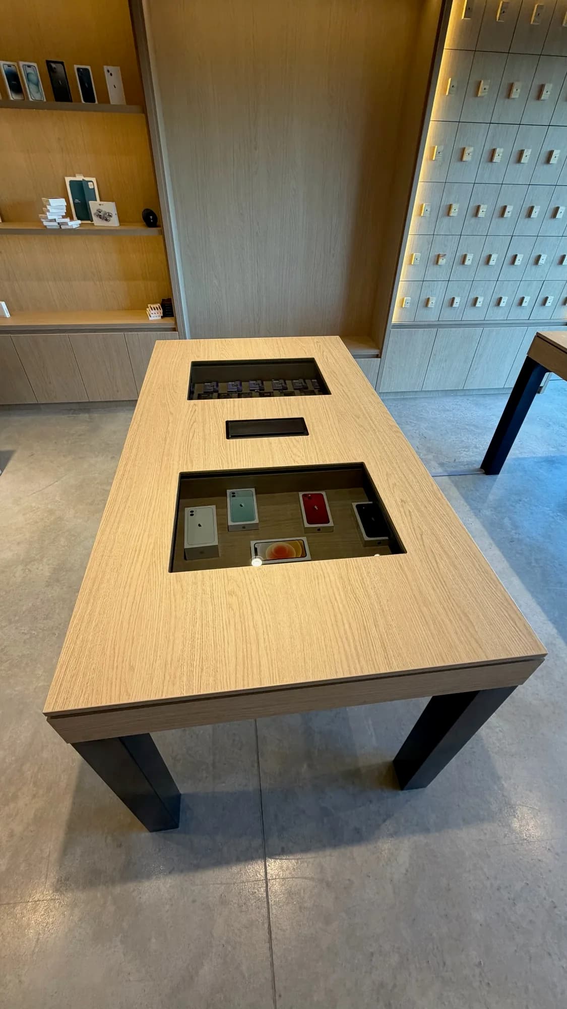 Mesa de exhibición en chapa de roble con patas negras y dos huecos vidriados que muestran teléfonos, frente a estantes y panel de casilleros iluminados; toma cenital en un local