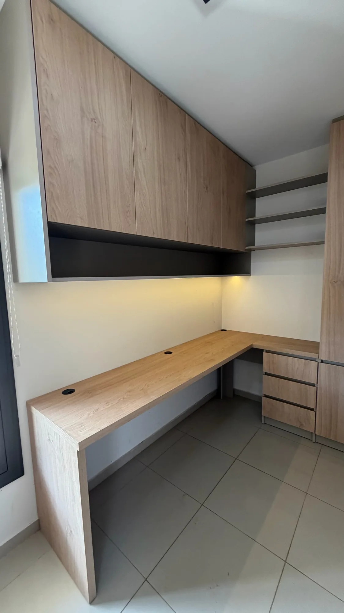 Escritorio en L de tapa símil roble con pasacables y cajonera, bajo alacenas altas con luz LED cálida debajo; toma angular de gran angular