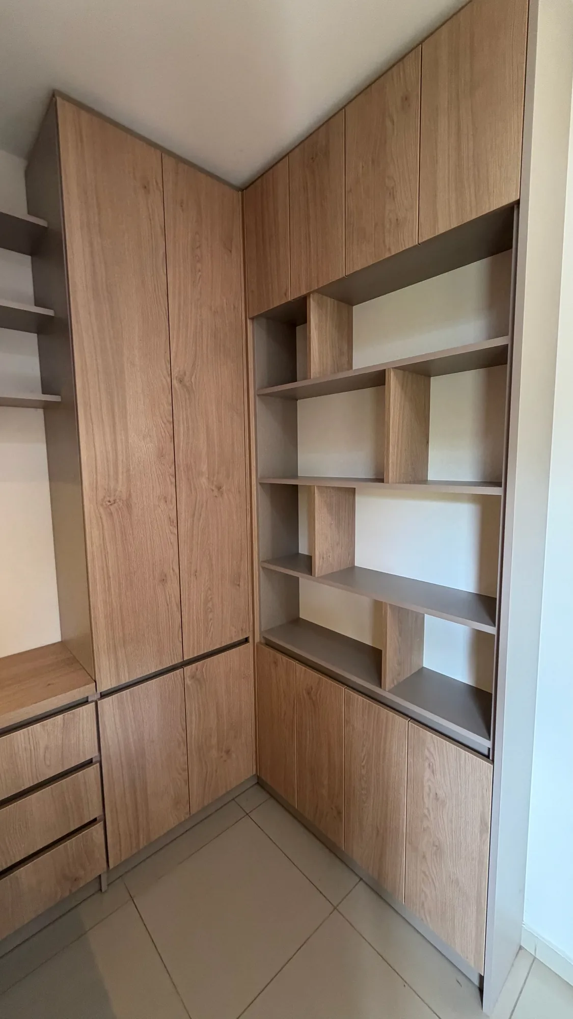 Rincón con placard alto, biblioteca de repisas abiertas y alacenas de puertas símil roble en melamina gris topo, con cajonera a la izquierda; toma en diagonal desde la esquina