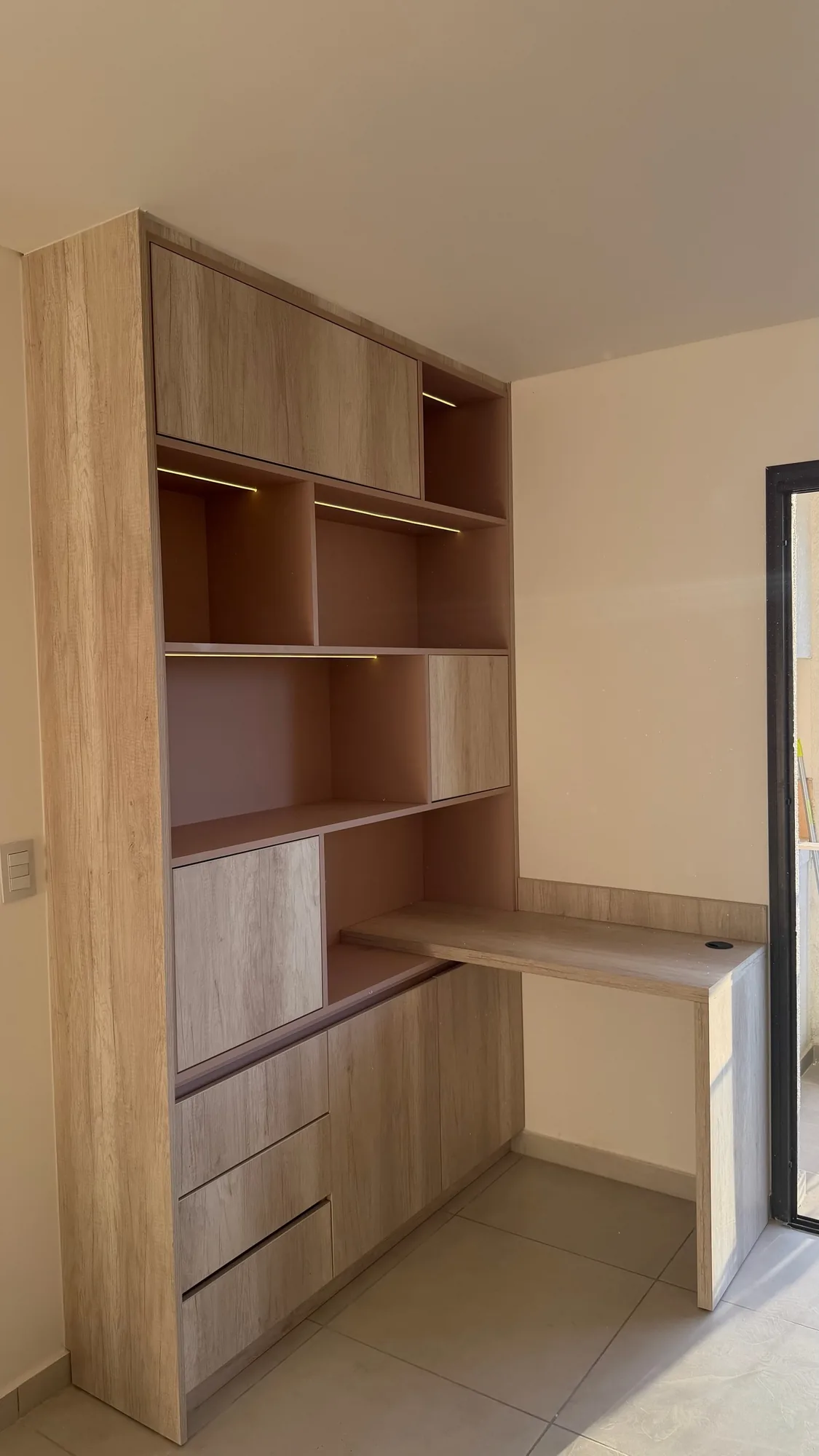 Biblioteca alta con escritorio integrado en melamina símil roble claro, con nichos con tira LED, cajones y puertas, junto a una abertura negra; toma oblicua completa