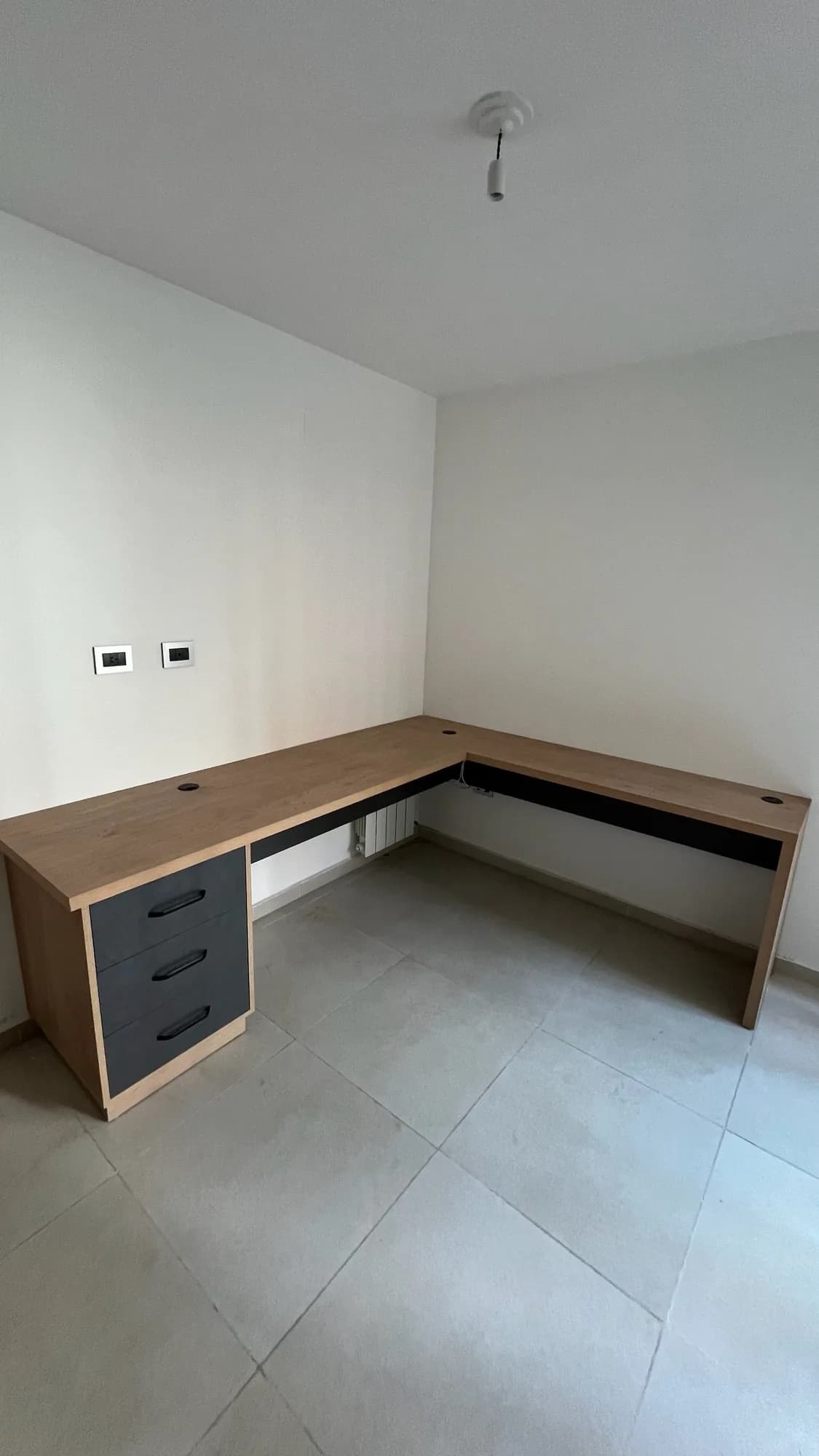 Escritorio en L en melamina símil madera con cajonera de tres cajones gris antracita y faldón oscuro, pasacables y enchufes en la pared; toma frontal vertical del rincón