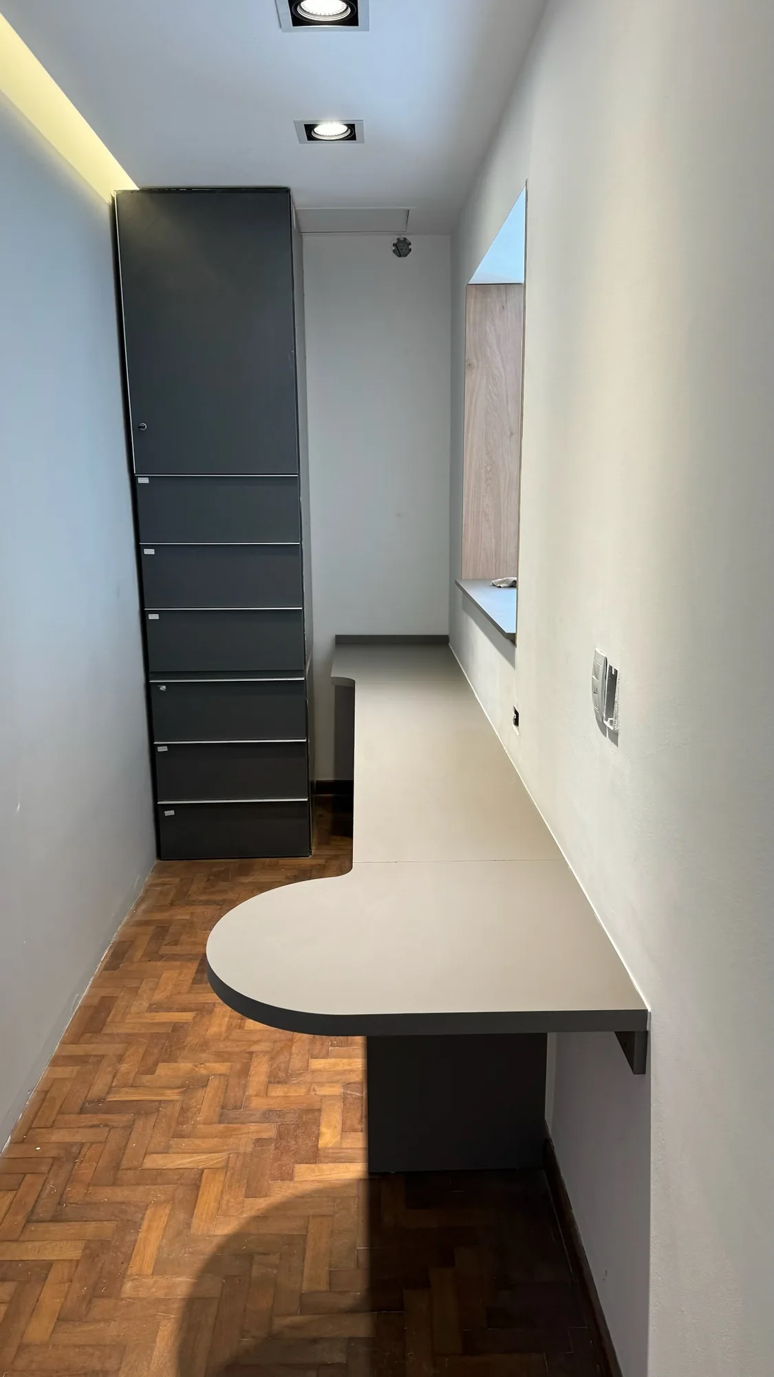 Escritorio largo gris con extremo curvo contra la pared, junto a un módulo alto gris oscuro con puerta y cajones, en una habitación con piso de parquet y spots empotrados