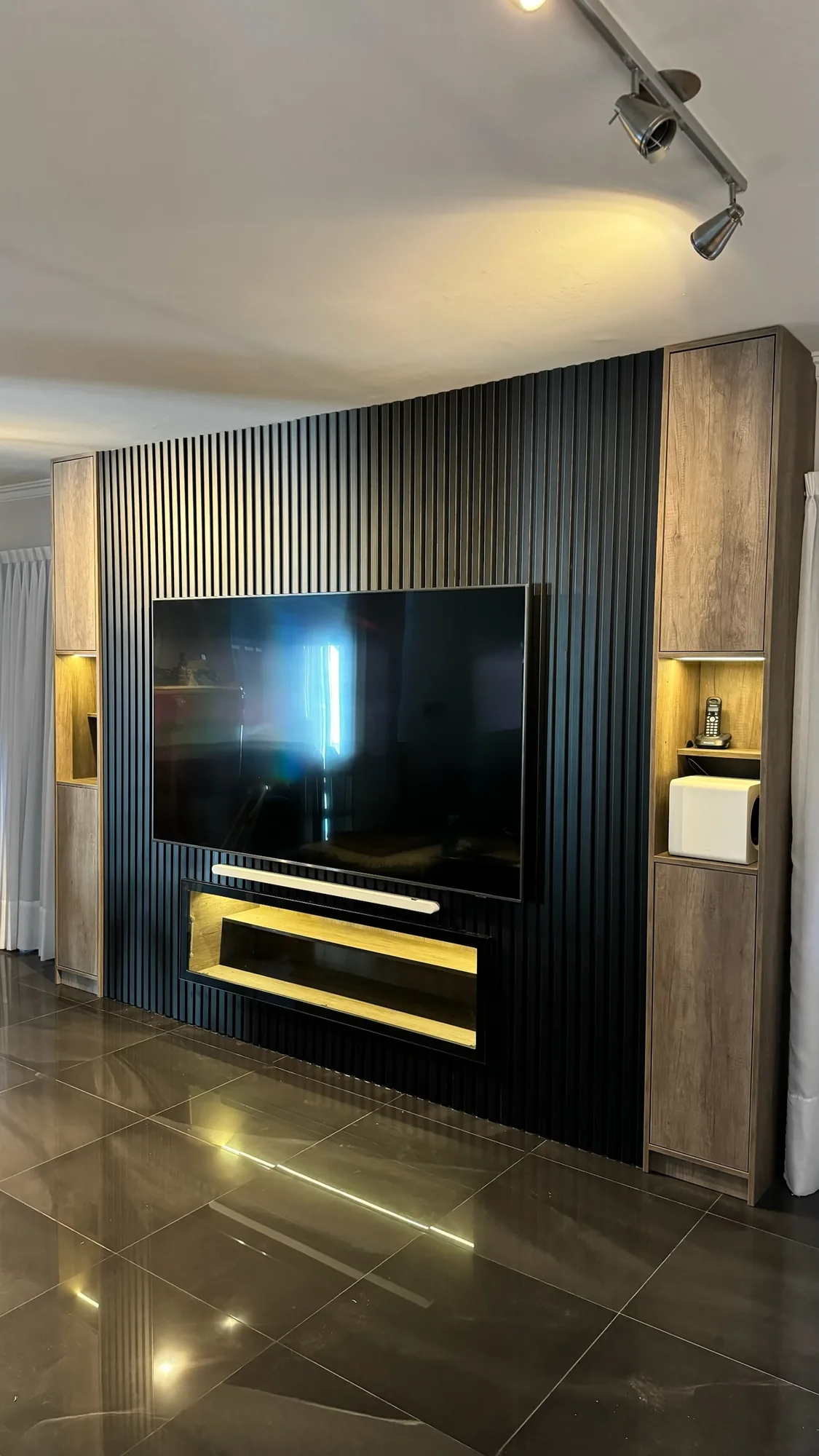 Mueble de living con panel de listones negros, TV colgada, repisa con nicho iluminado bajo la pantalla y dos columnas laterales de melamina símil madera con nichos con LED; toma frontal vertical