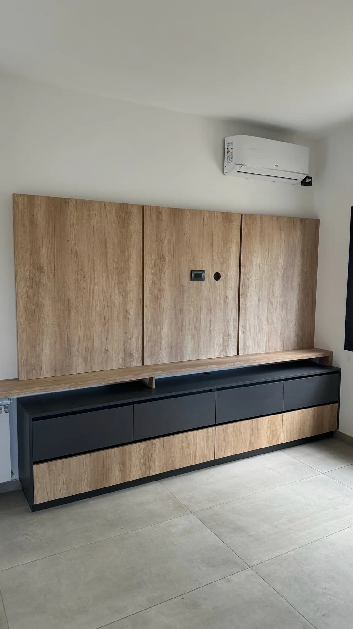 Panel de TV de tres módulos en melamina símil roble sobre rack flotante bajo con frentes antracita y madera, tapa de madera en voladizo y aire acondicionado arriba; toma frontal