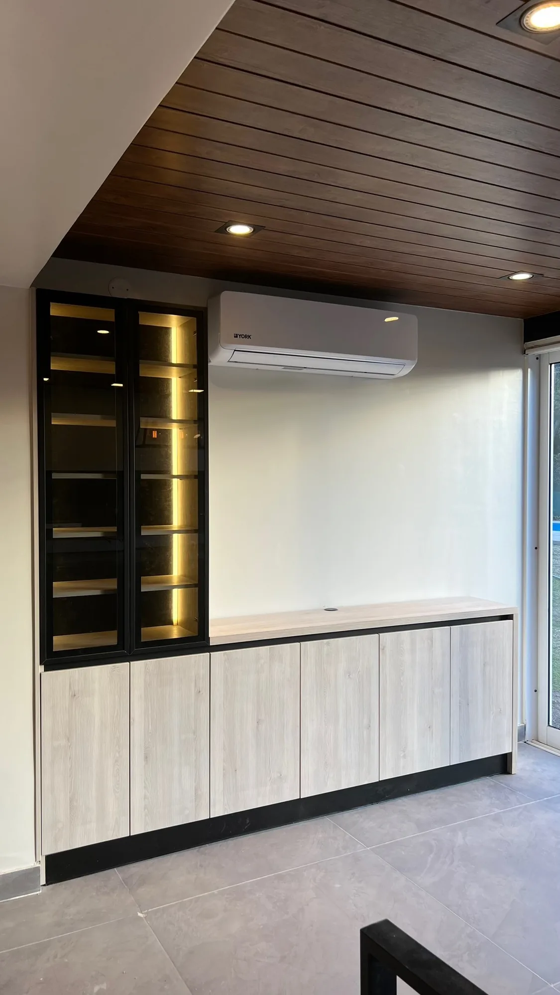 Aparador bajo con puertas de melamina clara y vitrina de aluminio negro con vidrio y estantes iluminados con LED, bajo un aire acondicionado y spots en el cielorraso de madera; toma en ángulo