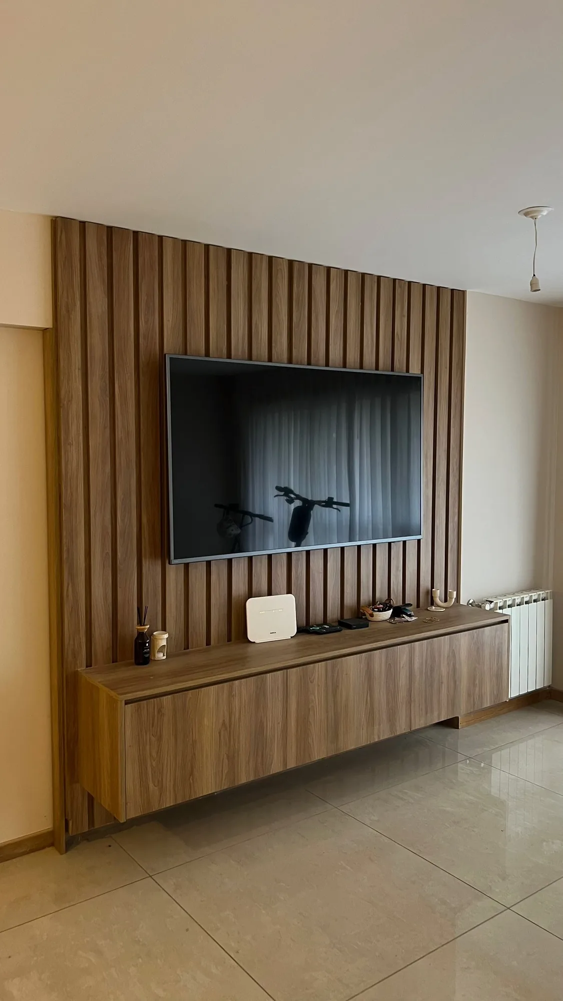 Panel de listones de madera con televisor y mueble de TV flotante de tres frentes en el mismo tono; toma frontal en un living con piso porcelanato y radiador al costado