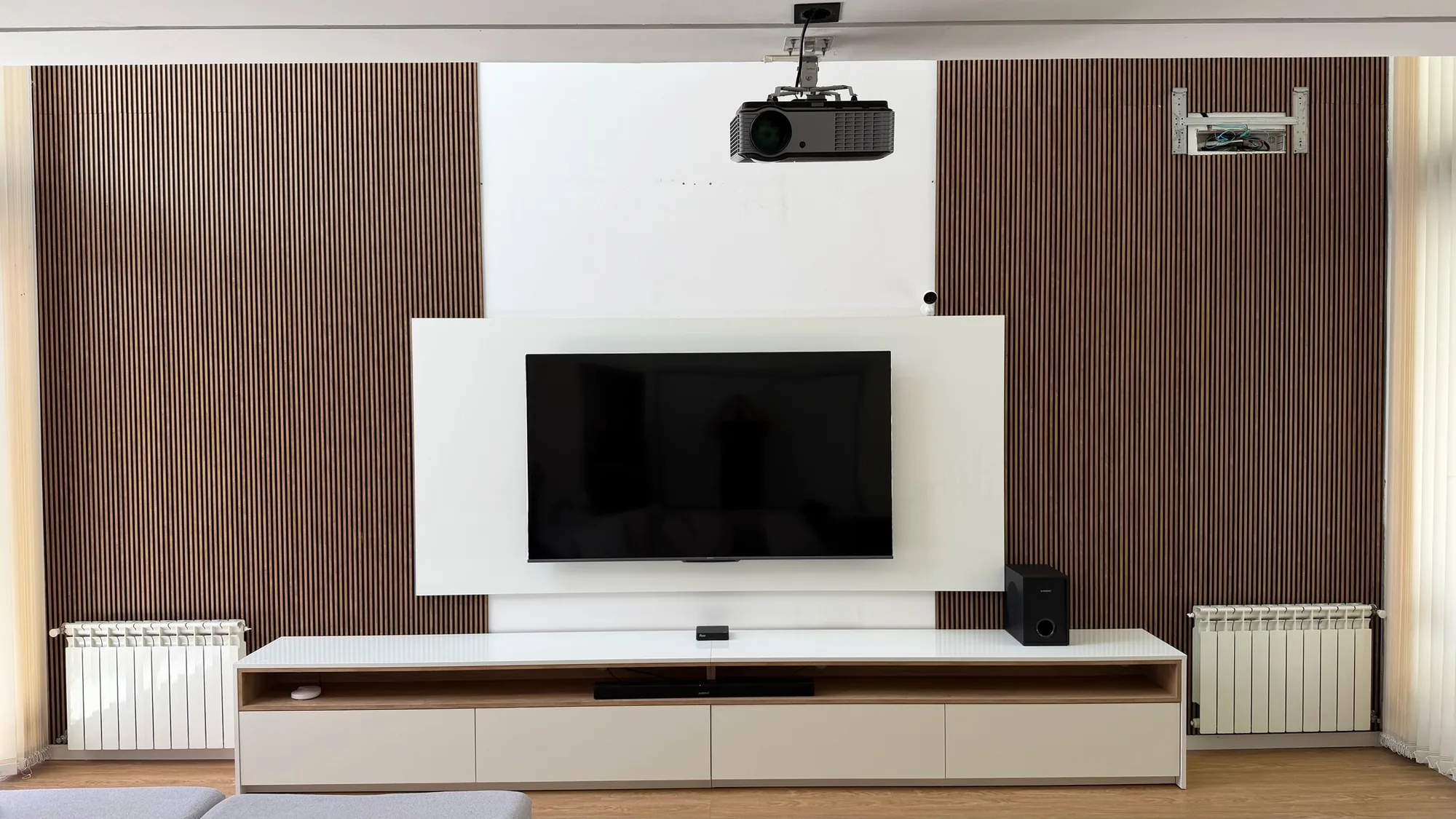 Rack de TV bajo en laca clara con detalle de madera y panel blanco flotante sobre pared de listones símil madera oscura; toma frontal y simétrica del living