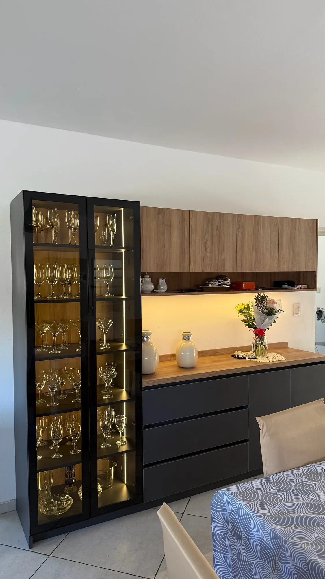 Vitrina alta de vidrio con tiras LED y copas, aparador gris grafito con mesada símil roble y alacenas altas con luz LED cálida; toma en diagonal desde el comedor