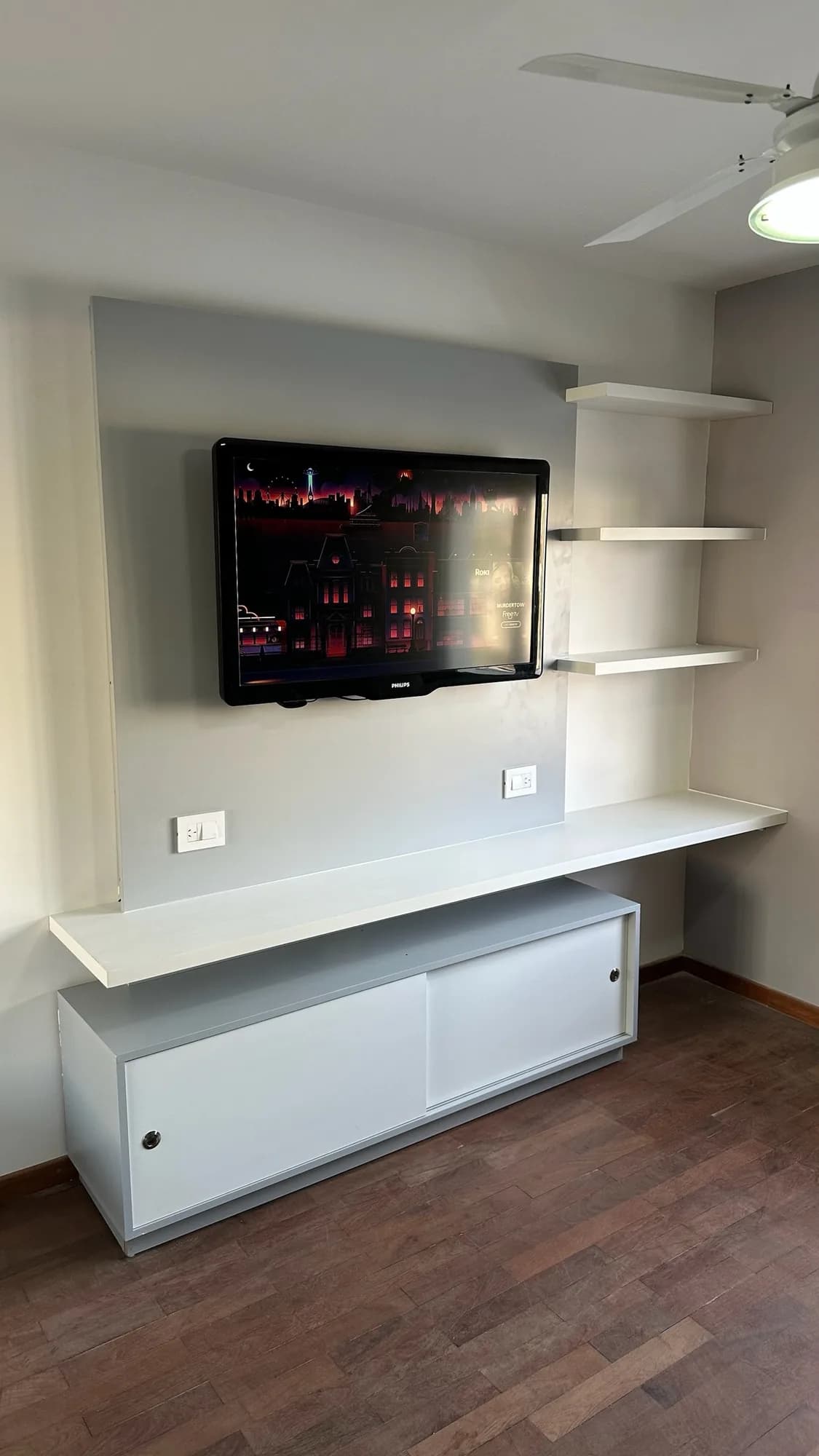 Panel gris con televisor, tres estantes flotantes blancos, repisa corrida y bajomesada con puertas corredizas blancas; toma frontal en un cuarto con piso de parquet