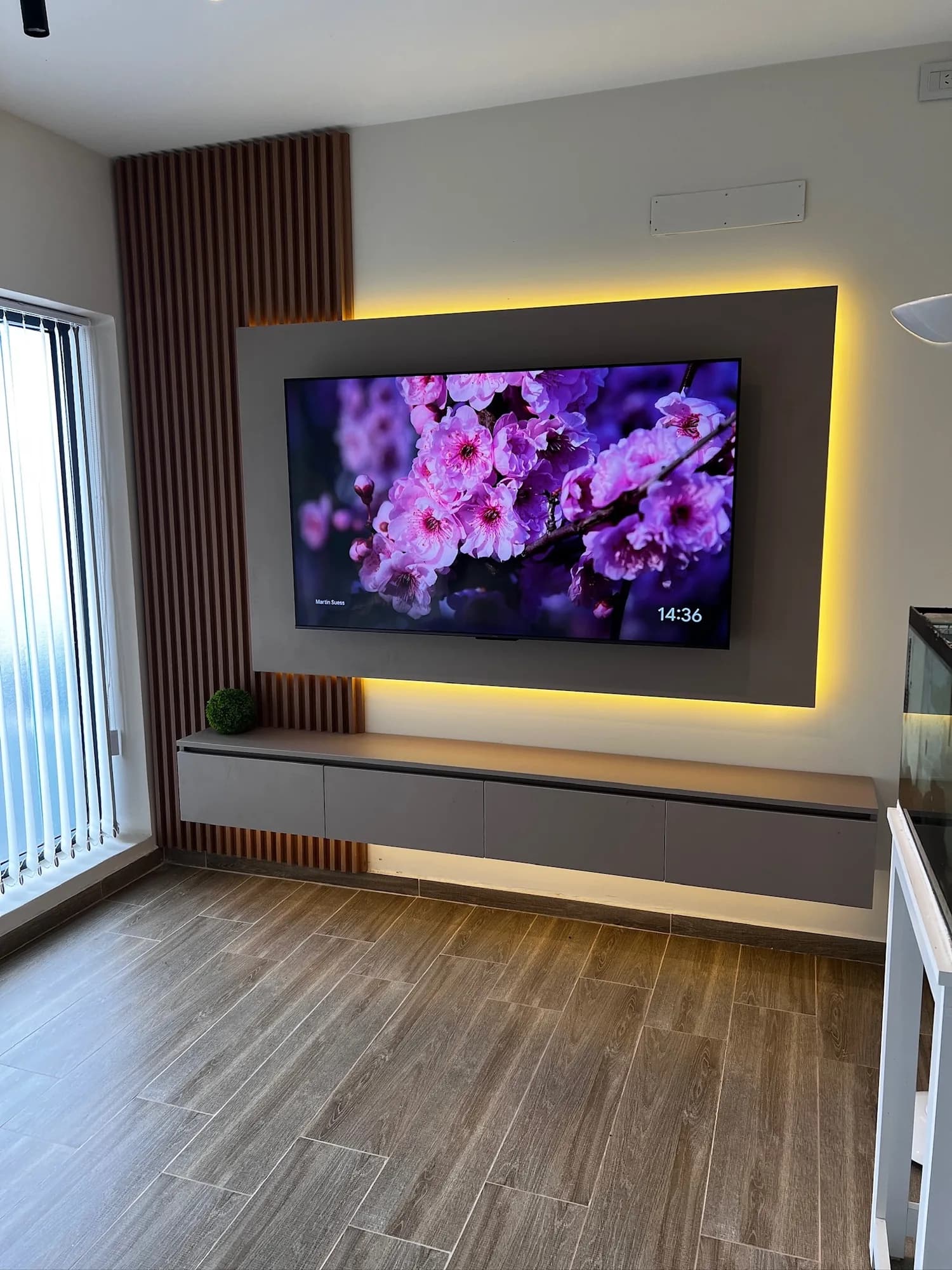 Panel de TV con marco gris y retroiluminación LED cálida, listones de madera a la izquierda y rack flotante de cuatro frentes; toma vertical en diagonal con ventanal a la izquierda