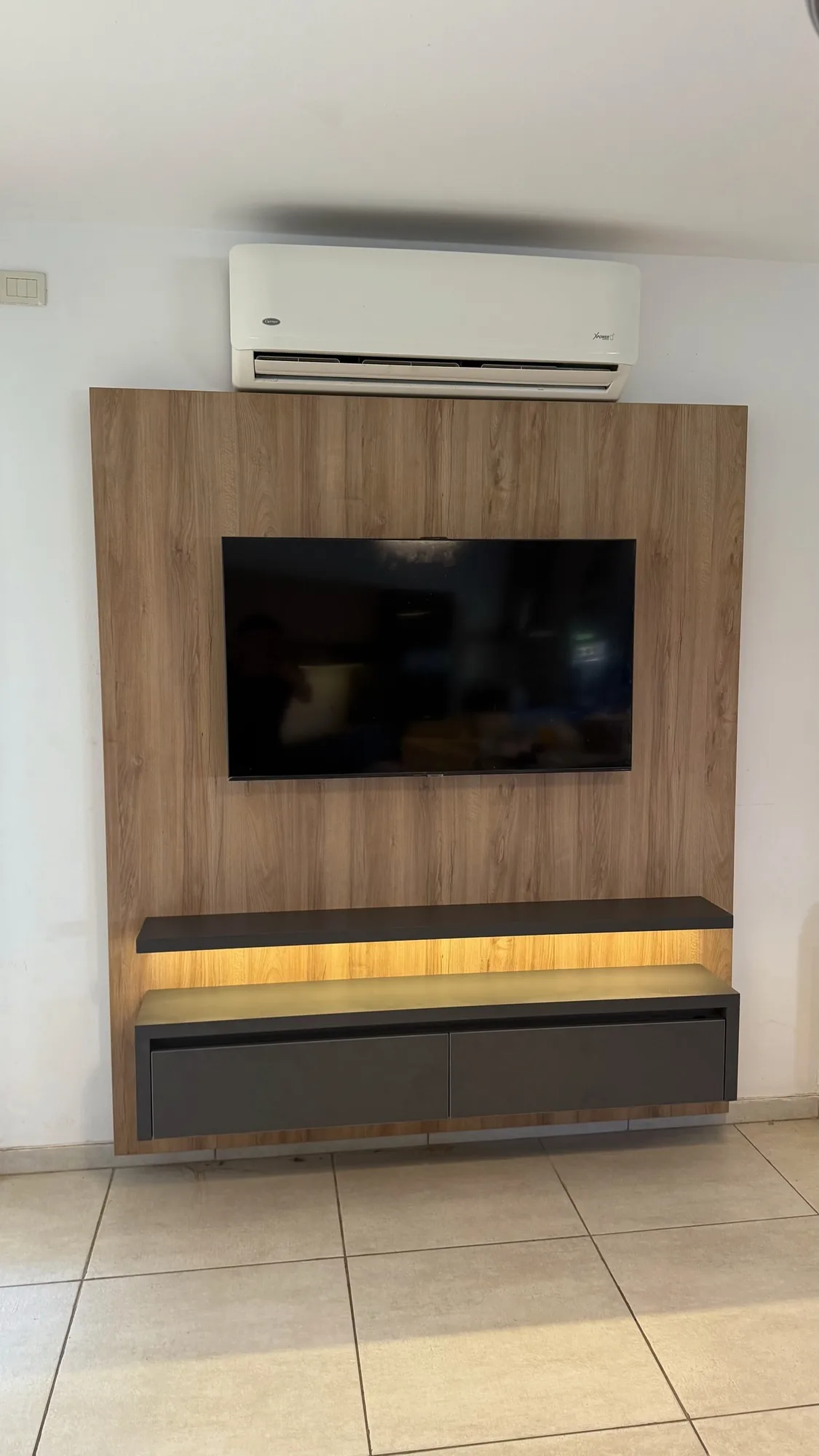Panel de TV en melamina símil roble con estante flotante oscuro, cajonera suspendida gris grafito y luz LED cálida detrás; toma frontal bajo un aire acondicionado