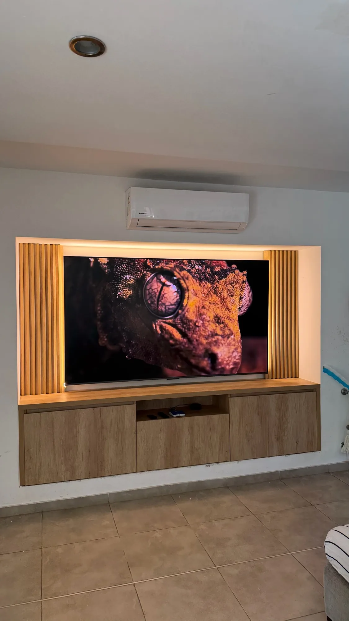 Panel de TV empotrado en nicho con listones de madera a los lados, retroiluminación LED cálida y rack flotante de tres puertas en melamina símil roble. Toma frontal con aire acondicionado sobre el nicho