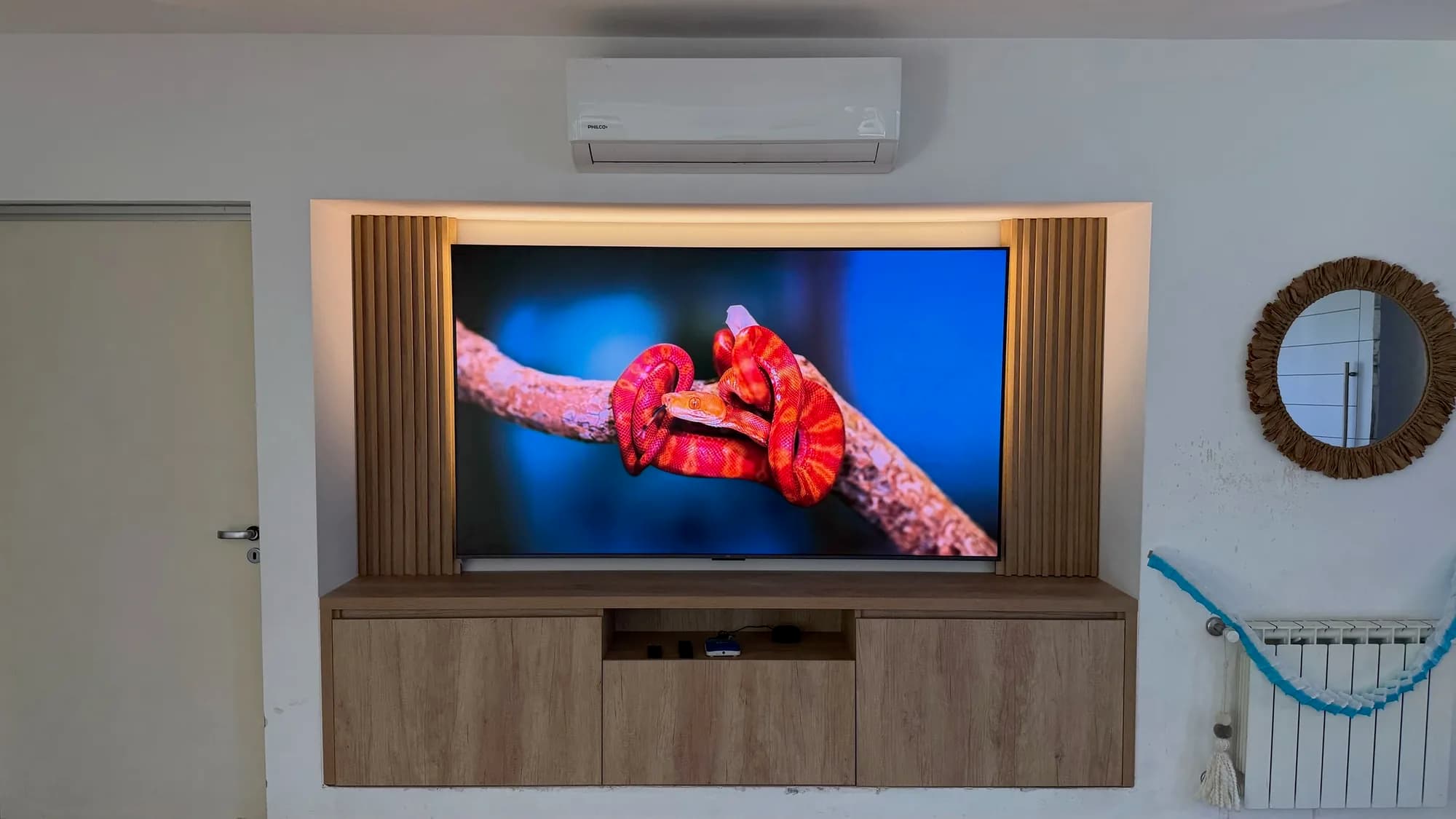 Panel de TV en nicho con listones de madera y retroiluminación LED cálida sobre rack flotante de tres puertas en melamina símil roble. Toma frontal general del living con aire acondicionado arriba